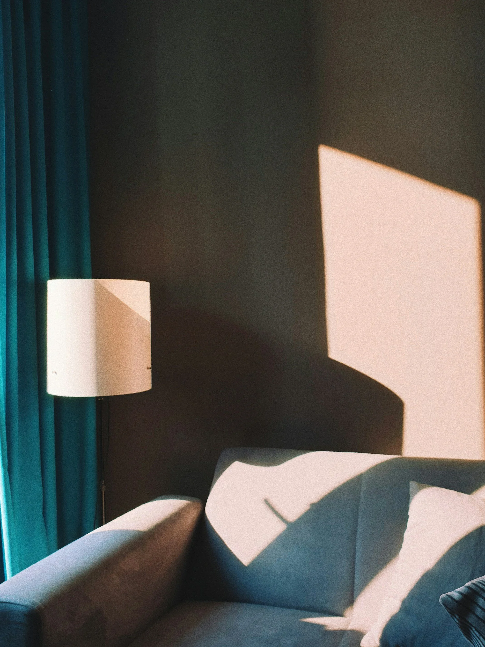 A cozy living room corner with a beige couch, a white throw pillow, teal curtains, a standing white lampshade, and sunlight casting geometric shadows on a dark wall.