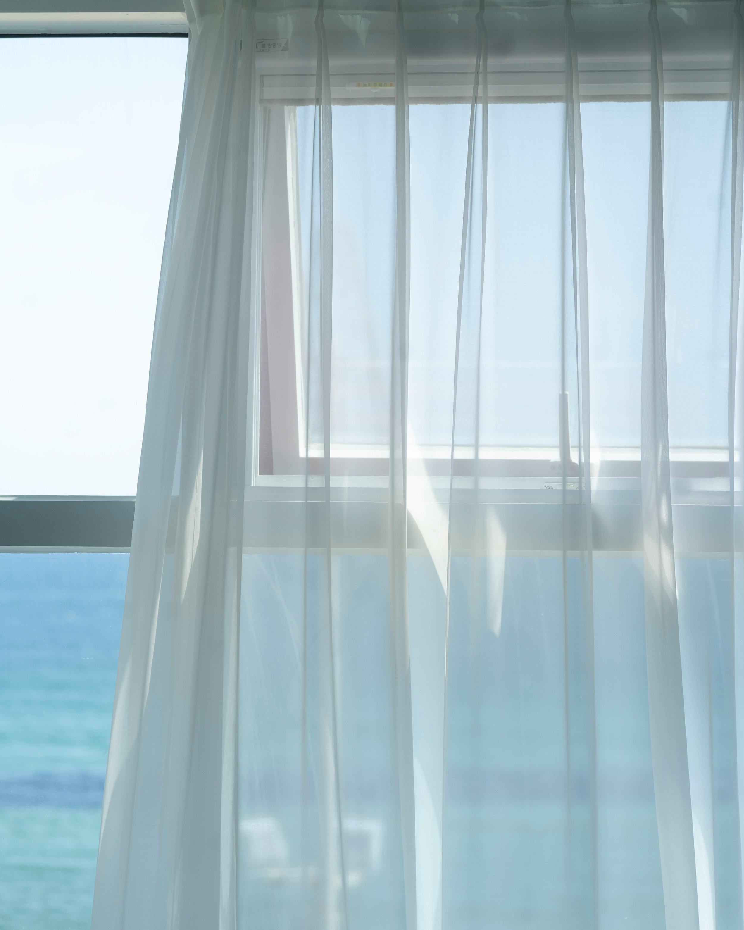 A sheer white curtain partially drawn back over a window with a view of the ocean and blue sky outside.