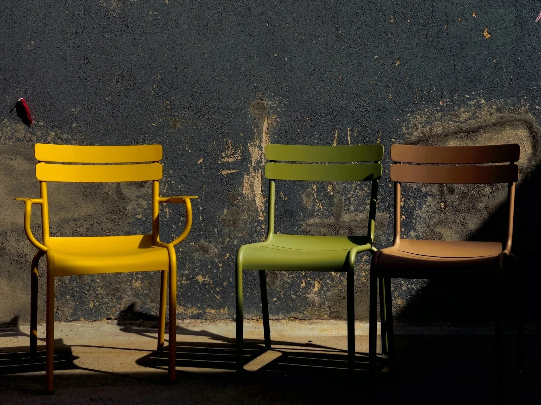 Three colorful chairs - yellow, green, and brown - are placed against a textured dark wall with sunlight casting shadows.