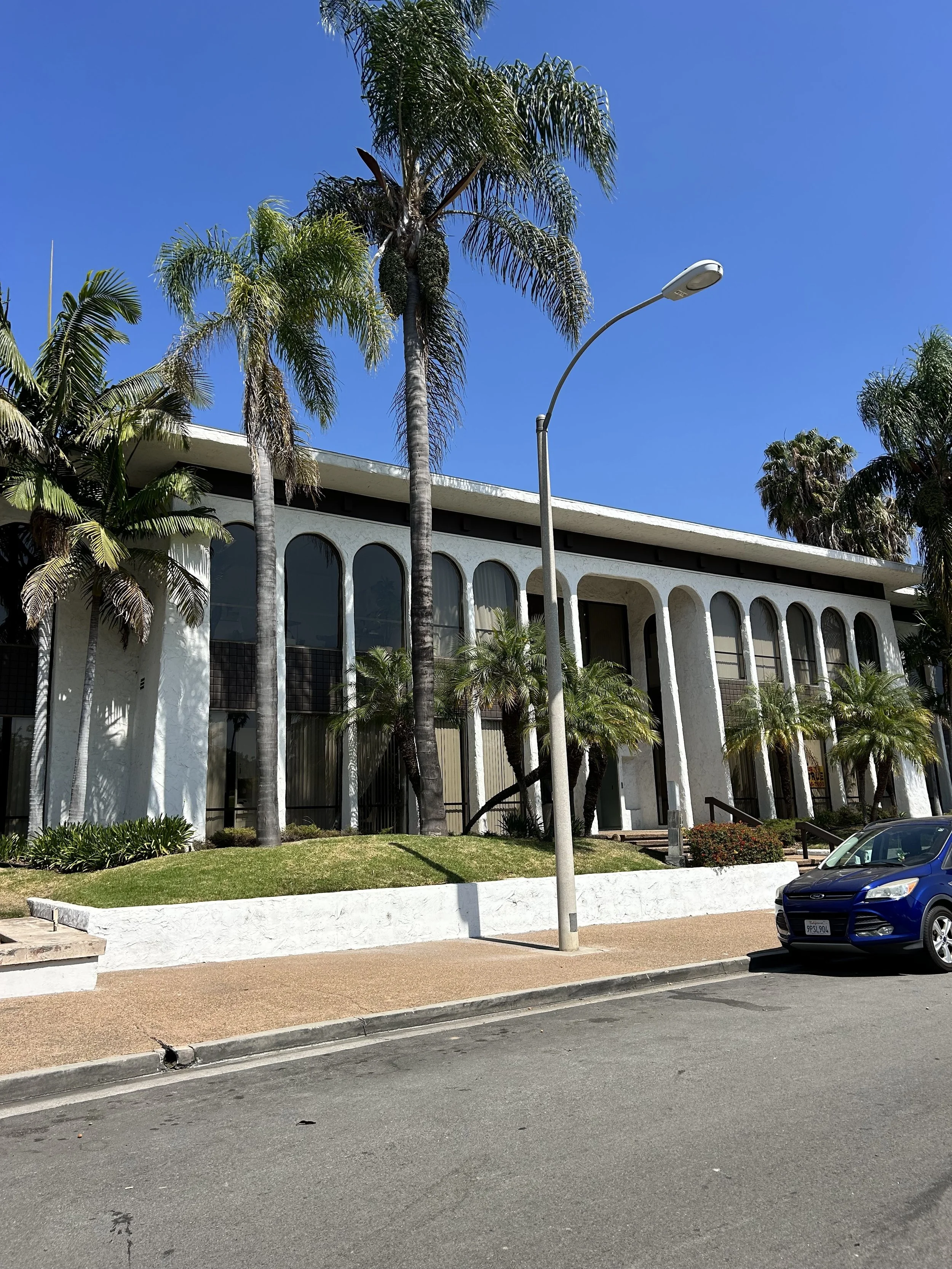 A building with white curved arches, surrounded by tall palm trees, under a bright blue sky. A street lamp and a dark blue car are visible in the foreground.