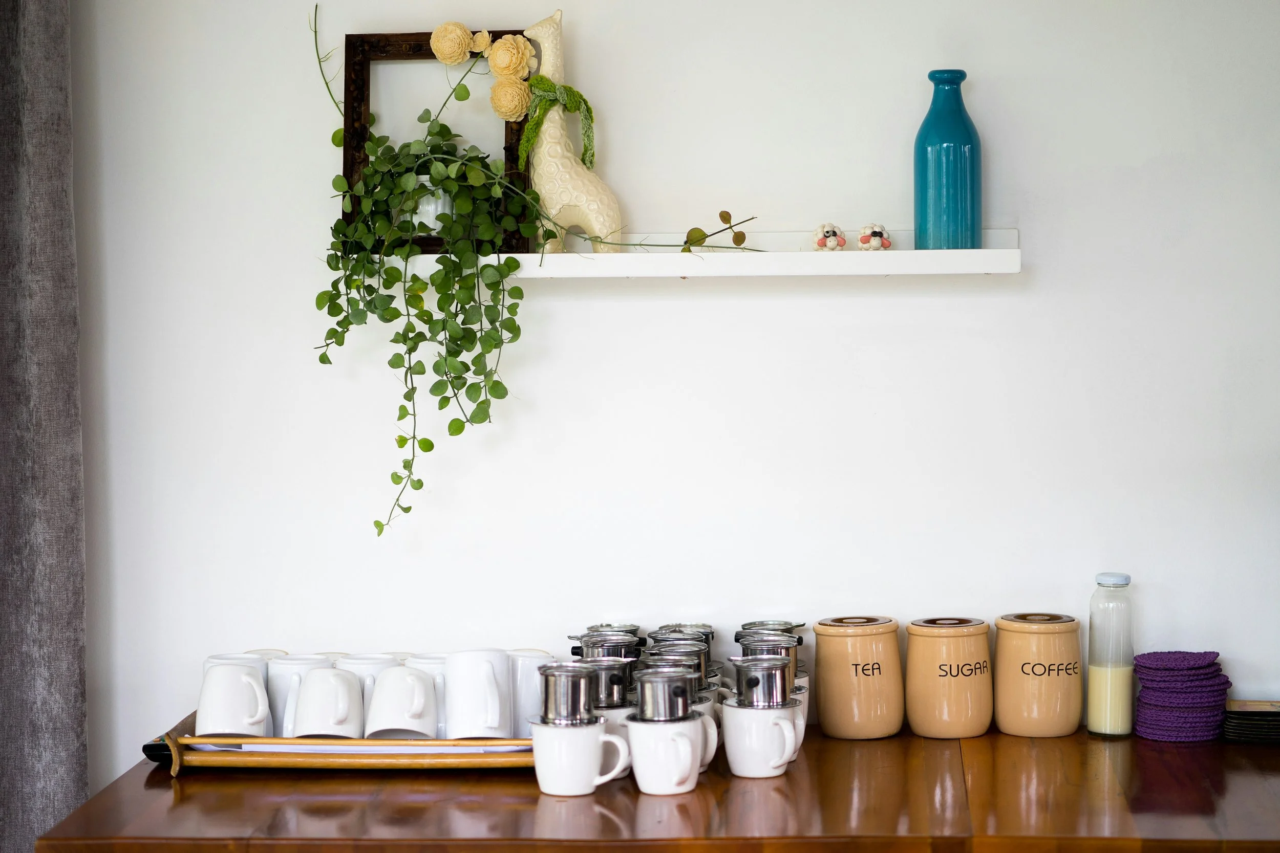 A row of white mugs and coffee canisters on a warm wooden counter, with a plant and framed art on a shelf above.