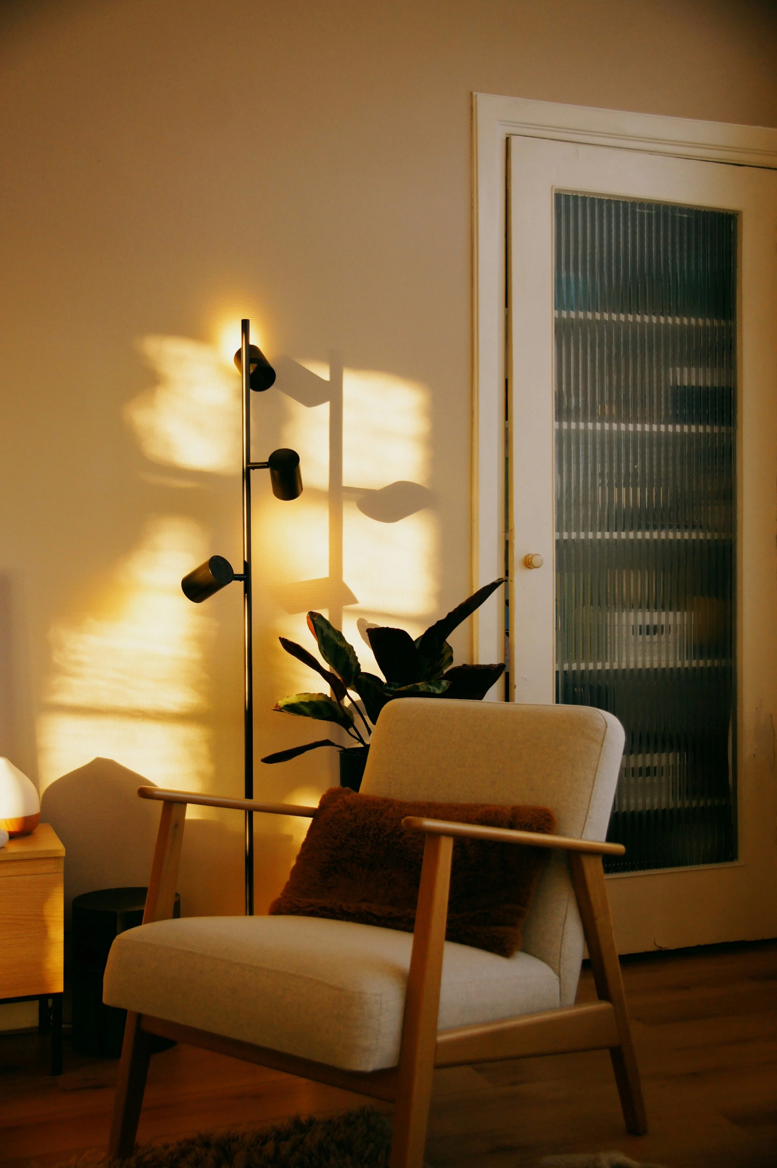A cozy living room corner with a beige armchair and a brown pillow, a black floor lamp casting shadows on the wall, a potted houseplant, a small side table, and a white door with a glass panel.