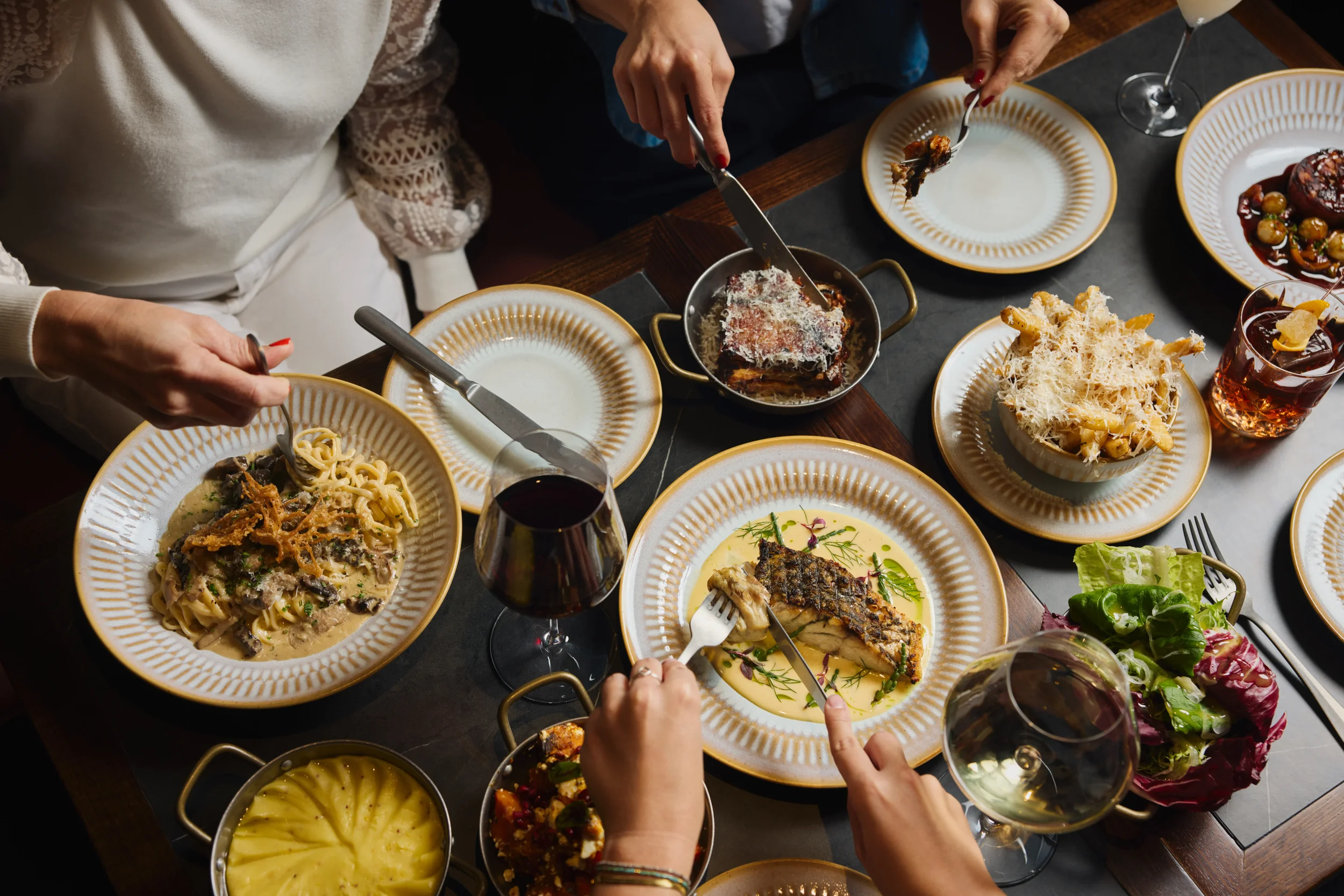 People sharing a meal with plates of pasta, fish, salad, and desserts on a dark dining table, with glasses of red and white wine.