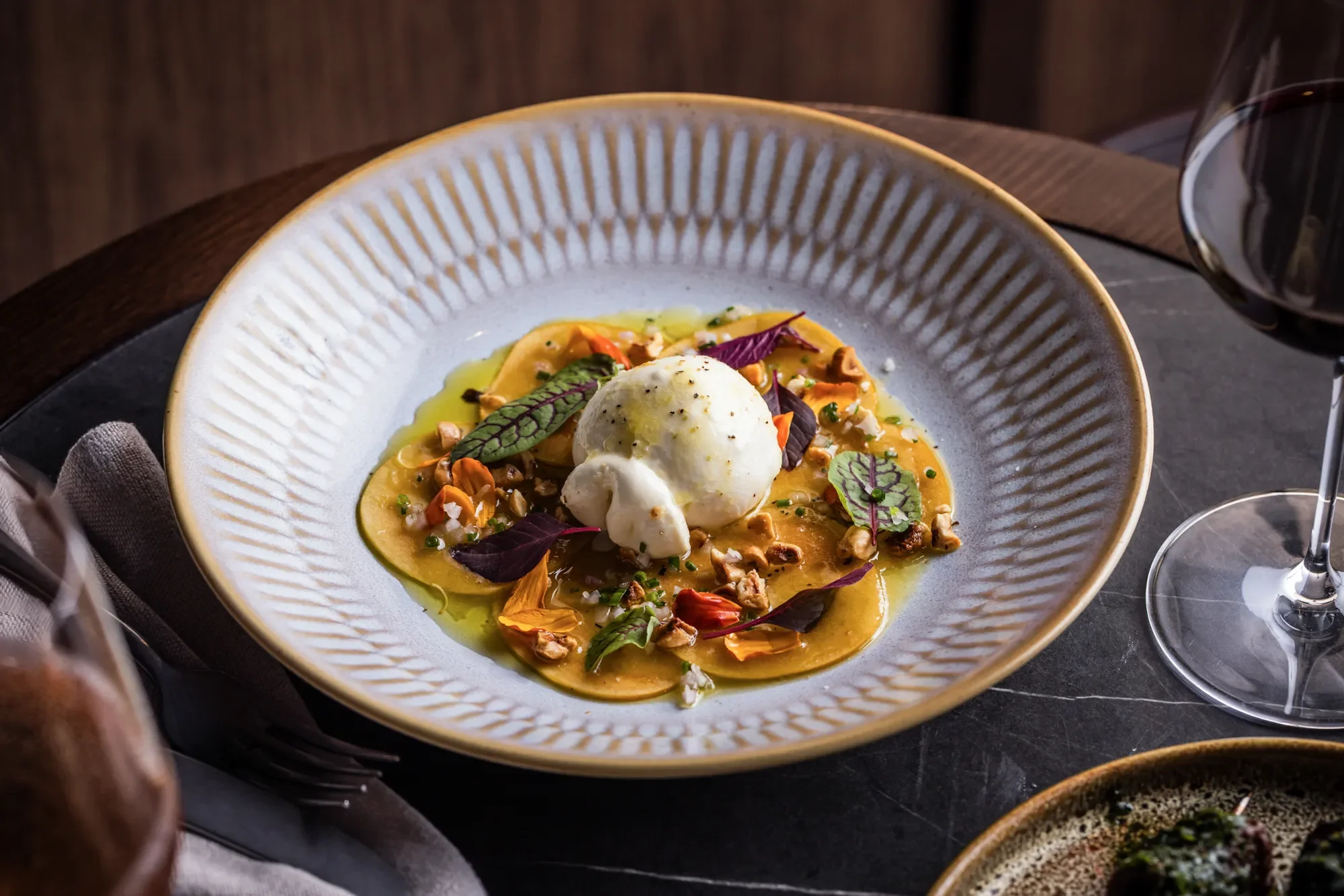 A gourmet dish with a scoop of white ice cream or sorbet, garnished with edible purple and green leaves, served on a yellow sauce with nuts and herbs on a white textured plate.