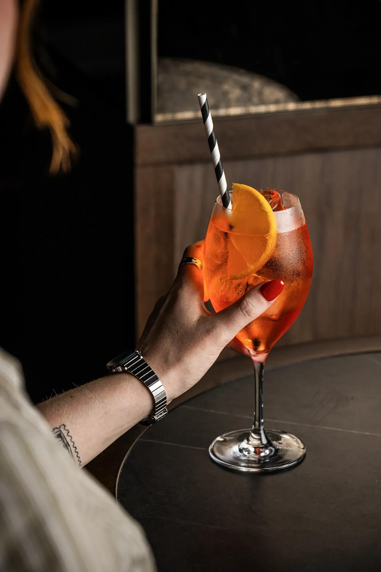 A person with a wristwatch and red nail polish holding a tall, elegant glass of orange-colored drink with ice, garnished with an orange slice and a black-and-white striped straw, against a dark background.