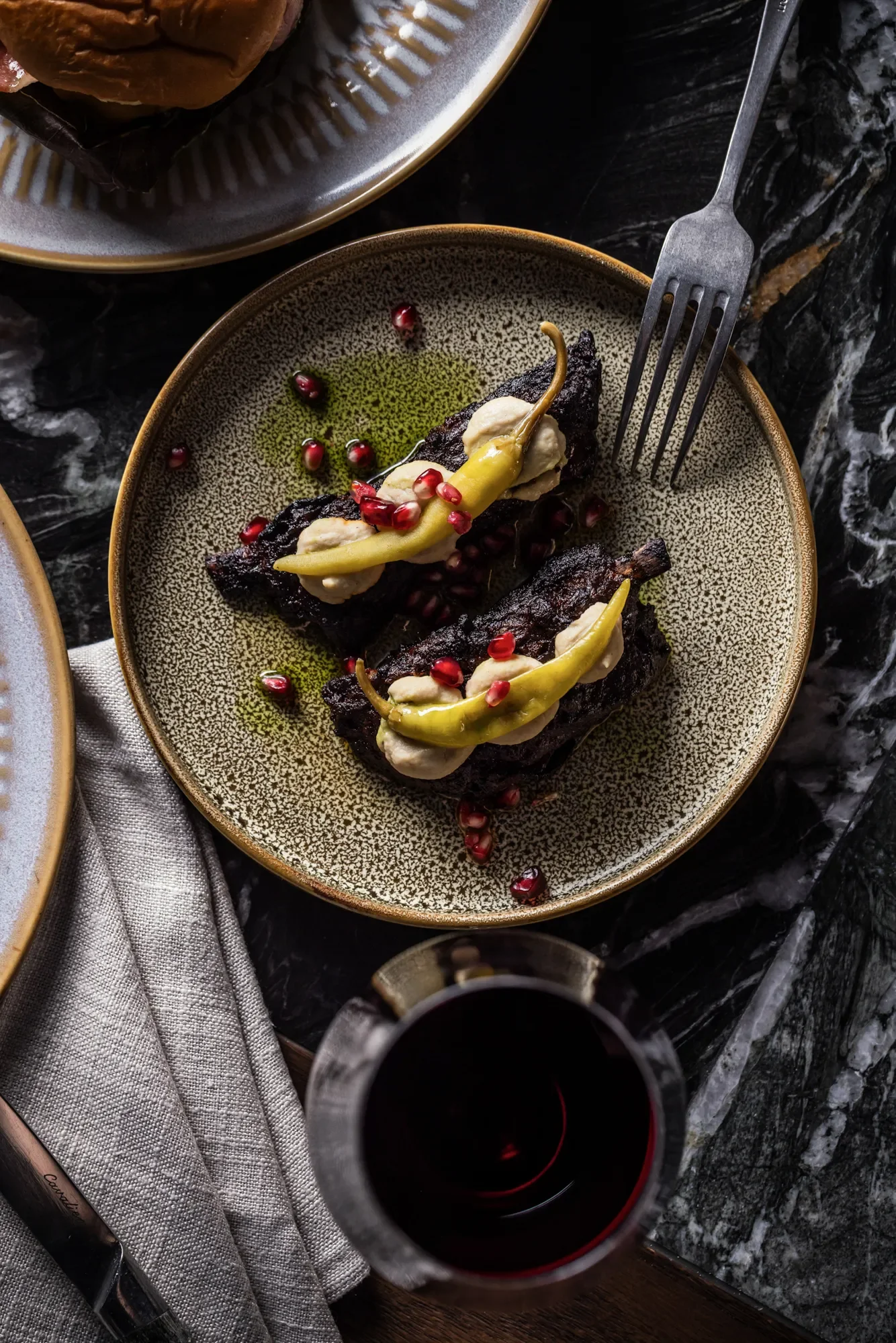 A plate of grilled meat topped with cheese, pickled peppers, and pomegranate seeds, with a glass of red wine nearby.