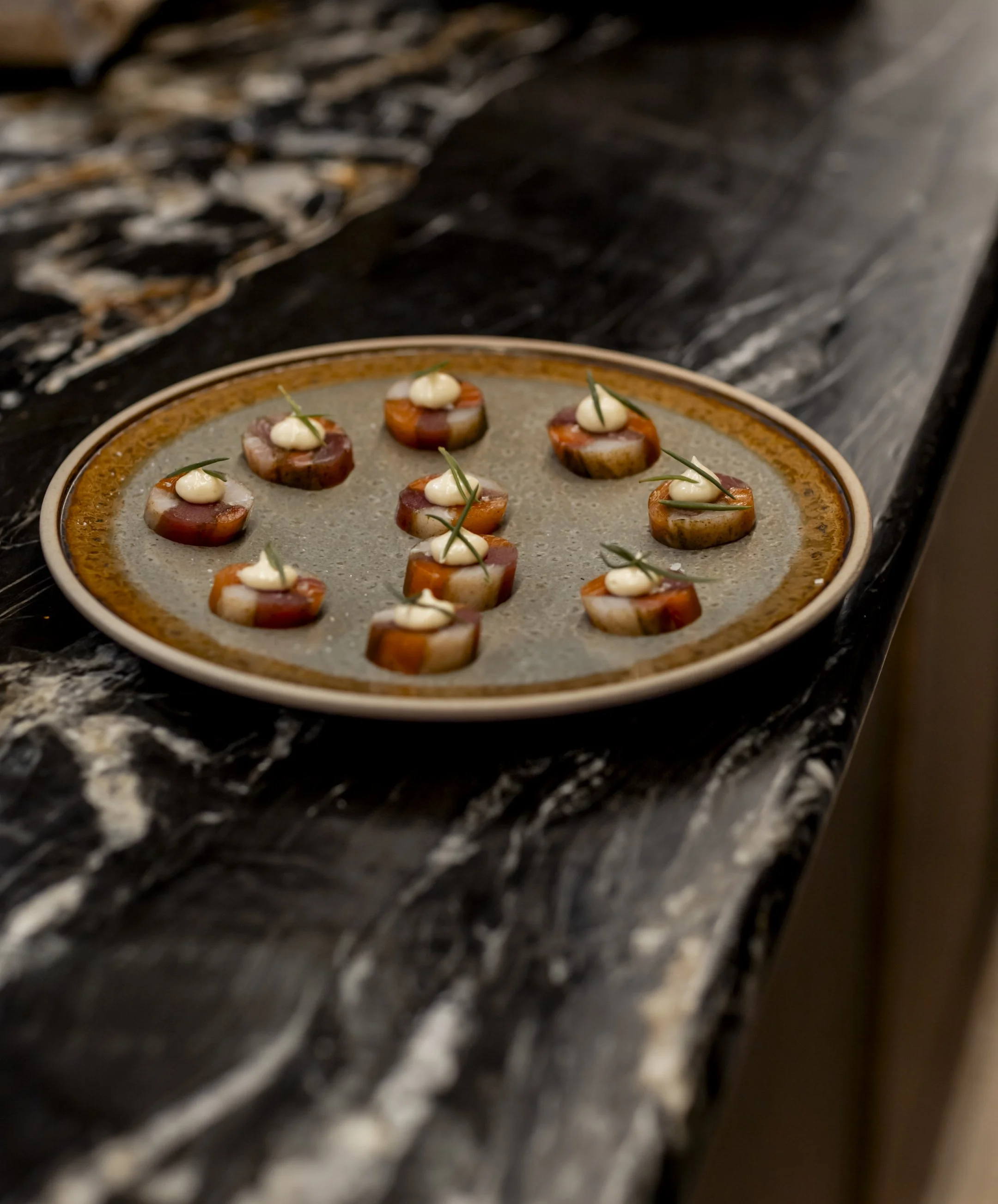 A round plate with smoked salmon canapés topped with dollops of cream and small green herbs, arranged on a dark marble countertop.