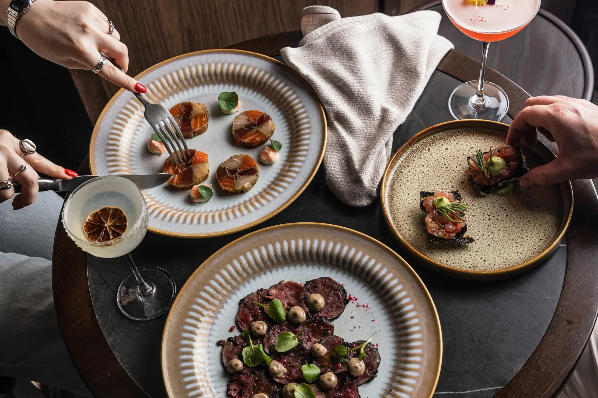 Gourmet dinner with three plates of food and two cocktails on a dark table. The dishes include sliced meat with sauce, a tartare topped with greens, and thinly sliced beef with pearl onions and microgreens. Two colorful cocktails are also on the tabl