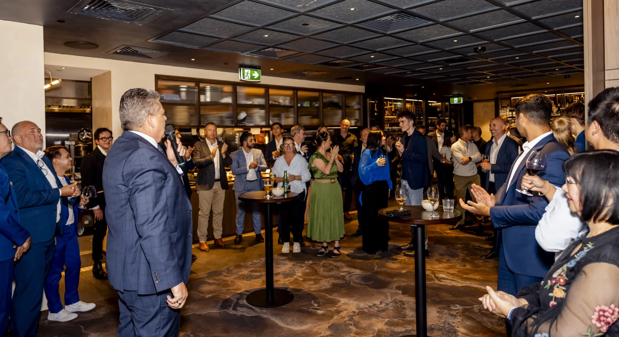 A man in a blue suit speaking into a microphone at a formal gathering, with guests clapping and holding drinks in a modern venue.