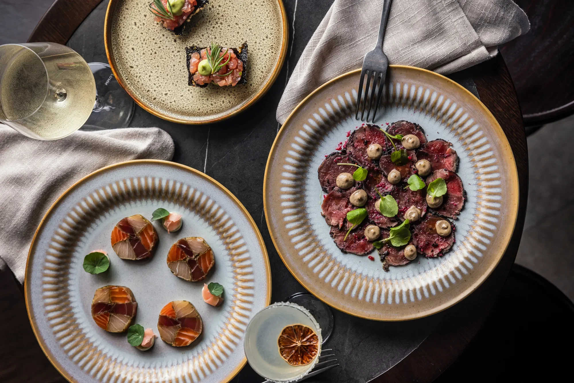 A top-down view of a table setting with three plates of food, a glass of white wine, and a cocktail. The plates feature thinly sliced cured meat garnished with greens, a plate of small layered bites topped with herbs, and another with tartare topped 