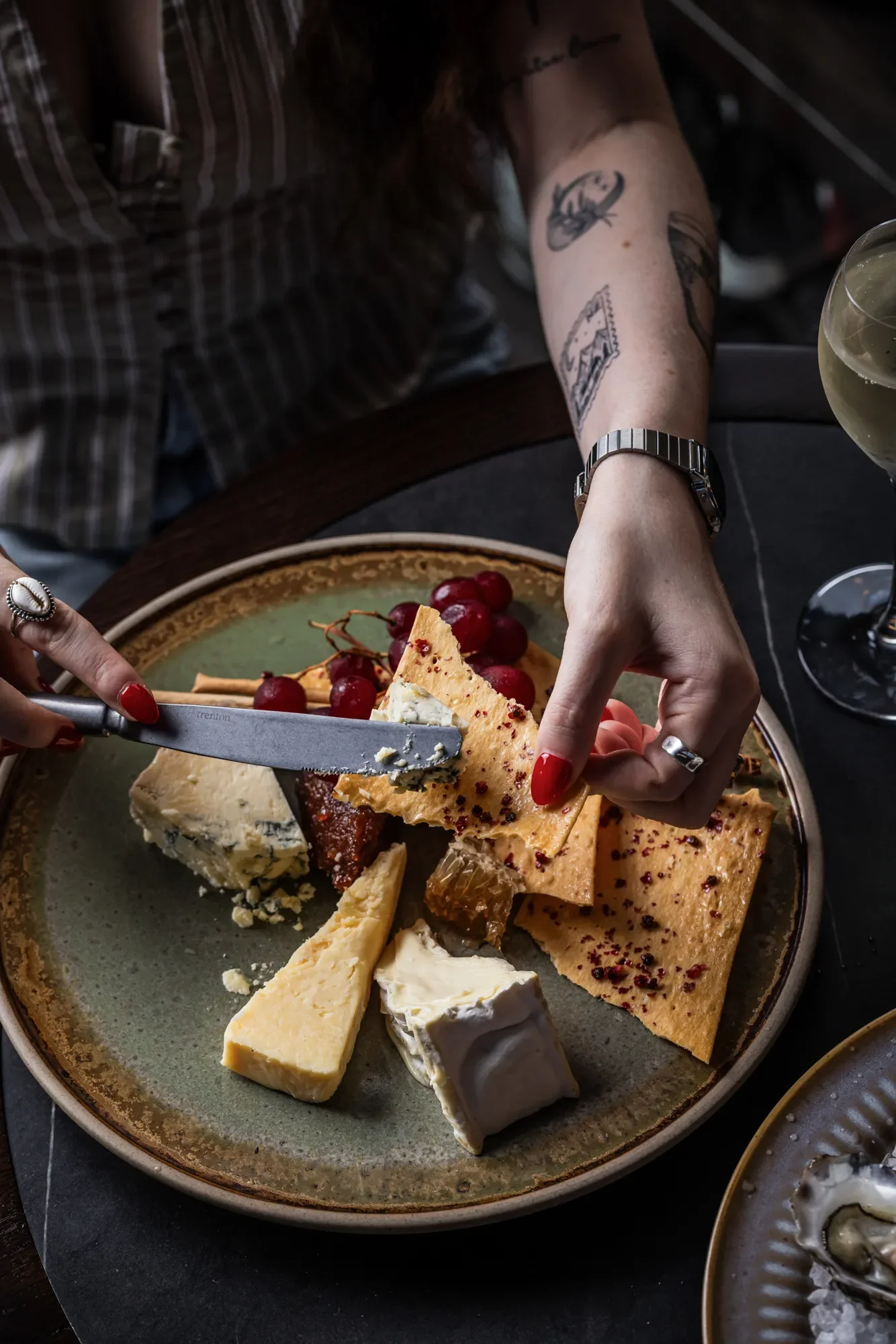 Person with tattoos and a watch slicing a cheese and cracker platter with grapes and a glass of white wine nearby.