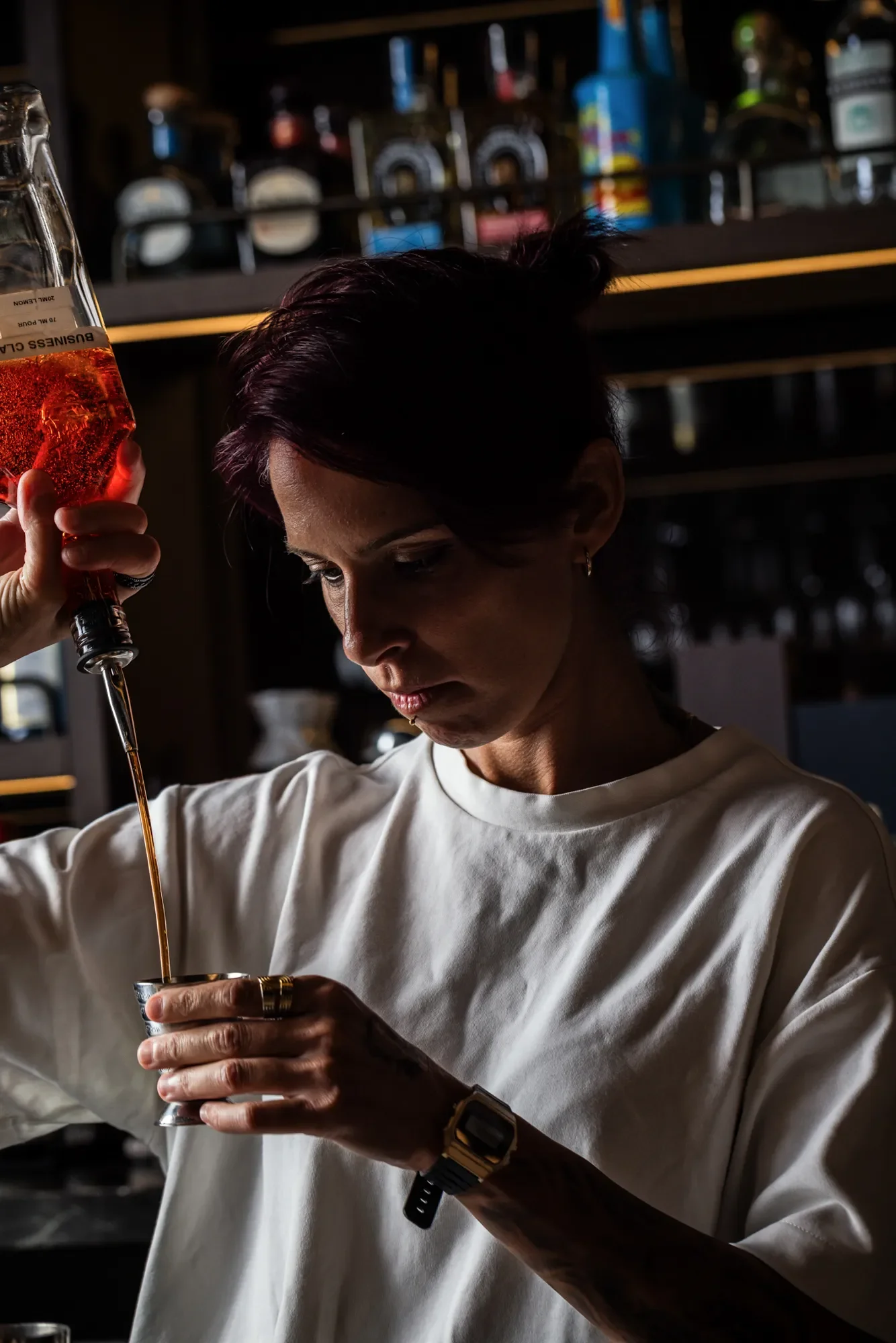 A woman behind the bar pouring alcohol from a bottle into a metal cup in a dimly lit bar.