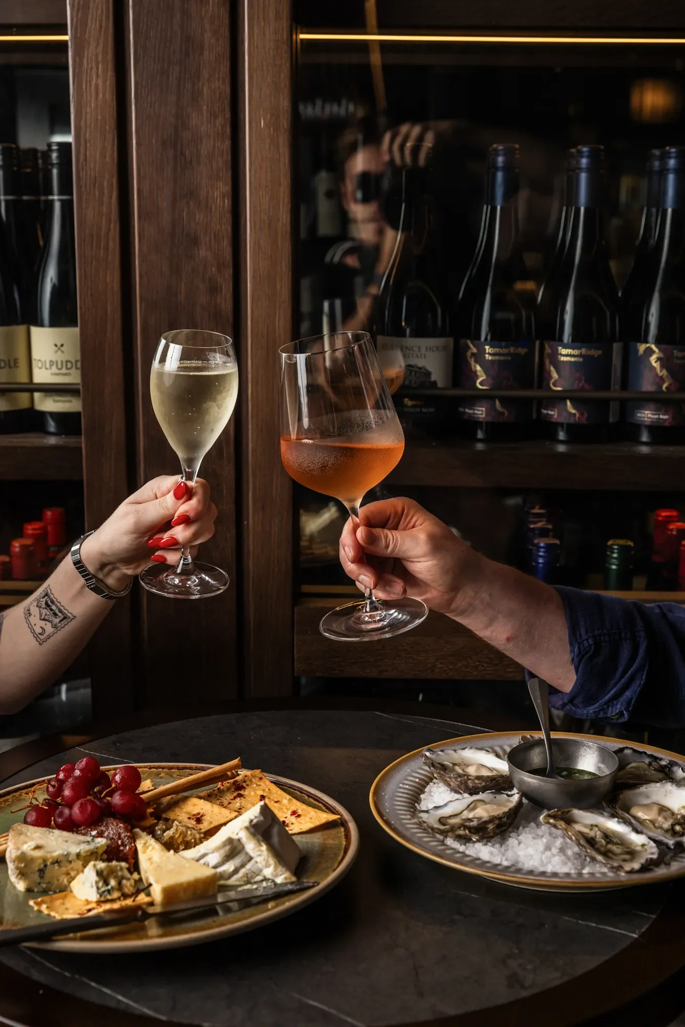 Two people clinking glasses of white wine and rosé wine at a cozy wine bar with cheese and oysters on the table.