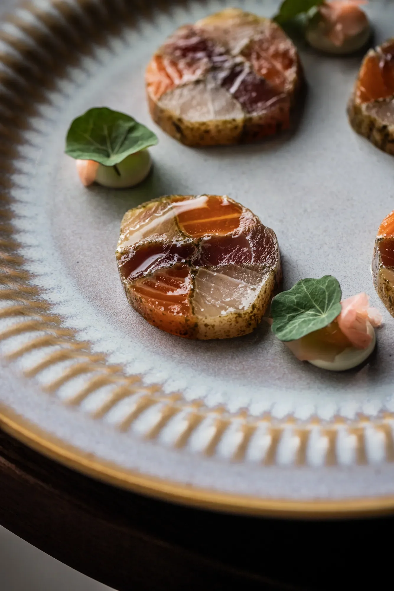 Close-up of a decorative plate with assorted savory jellies or aspics, garnished with small green leaves and pink edible flowers.