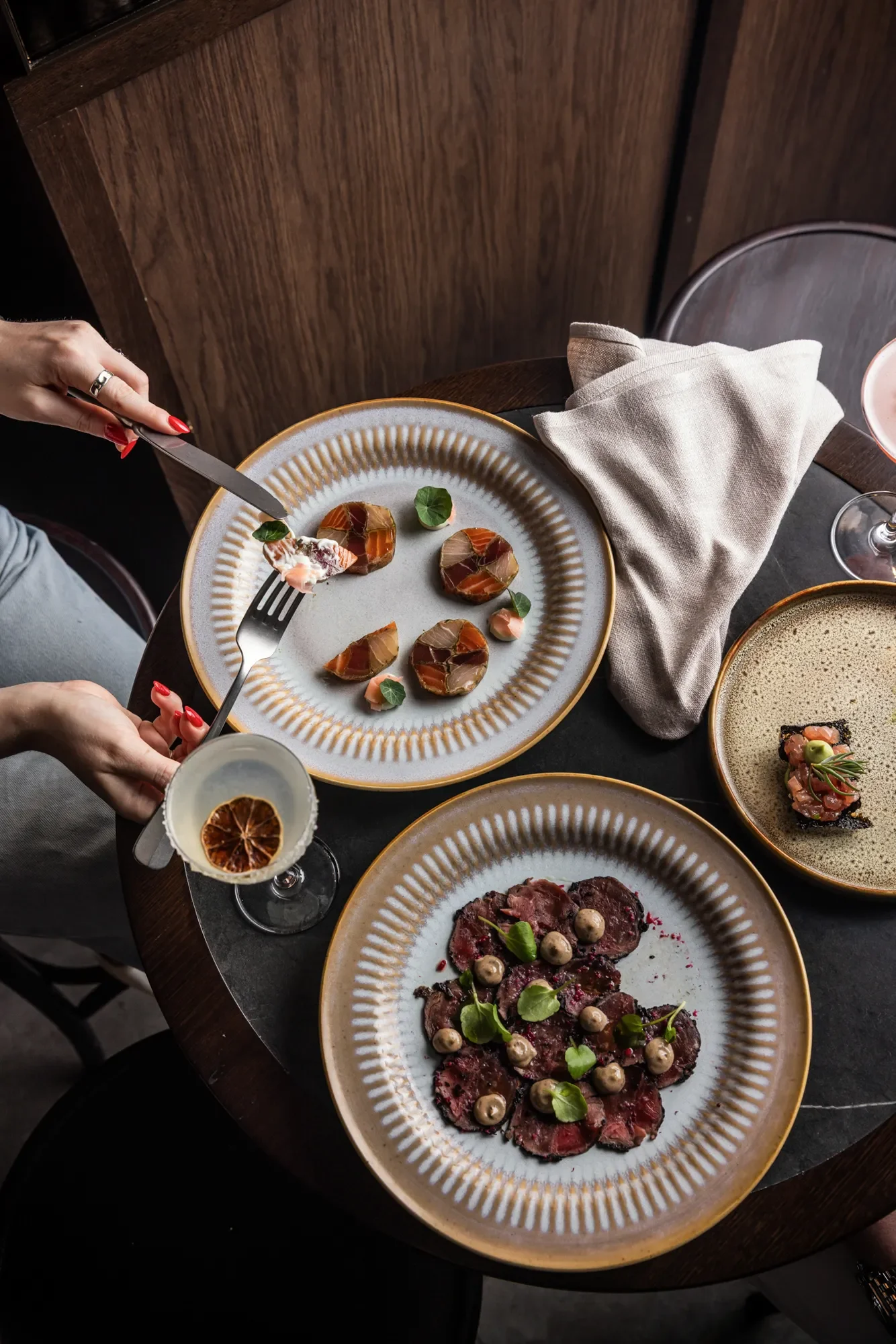 Overhead view of a table with three plates of gourmet appetizers, a glass of white wine with a dried citrus slice, a beige napkin, and a pink cocktail glass, set on a dark stone table with a wooden background.