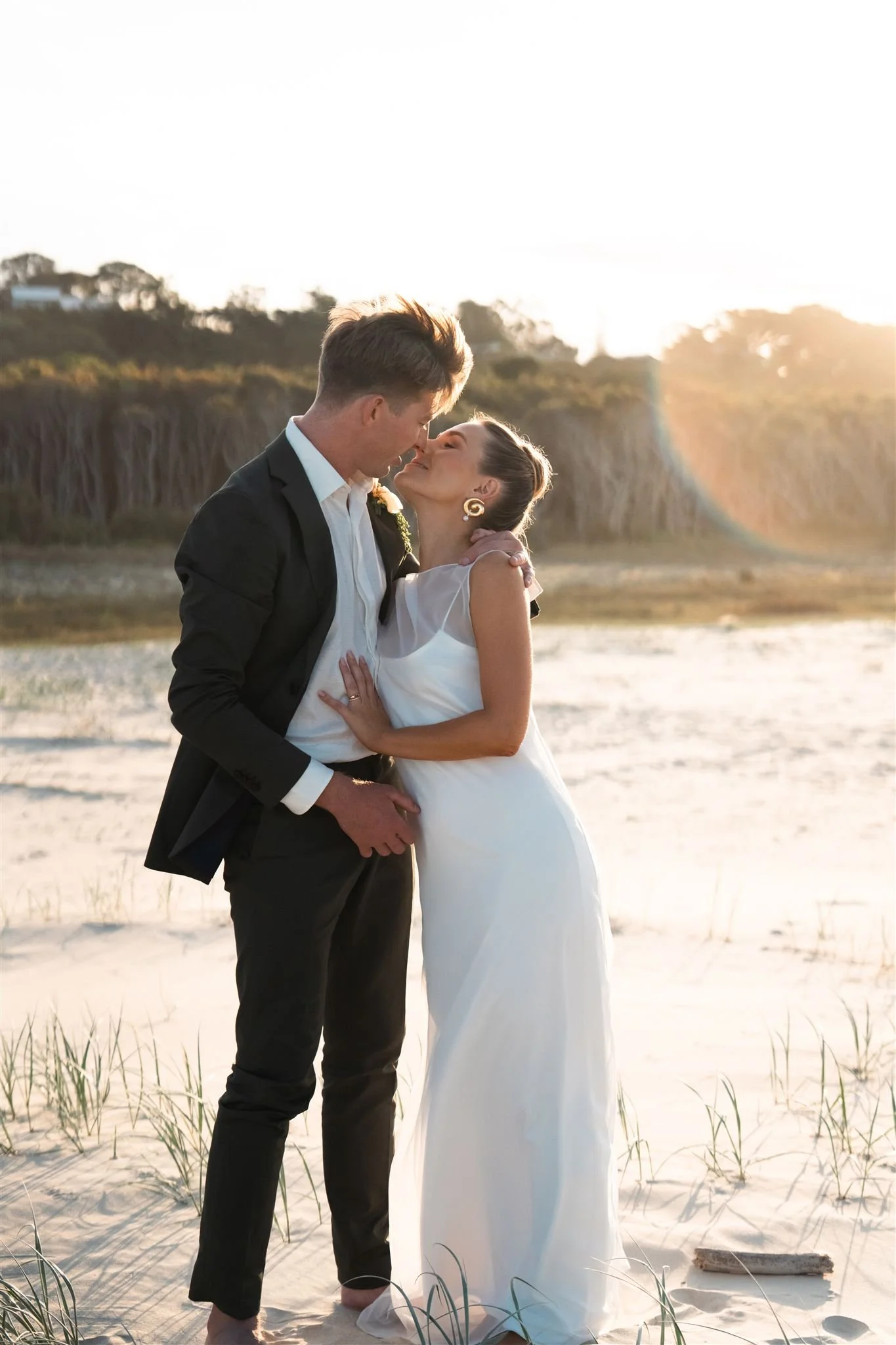 Beach wedding, bride and groom, A couple sharing a romantic moment on the beach during sunset, with the man in a black suit and the woman in a white dress, standing close and touching foreheads.