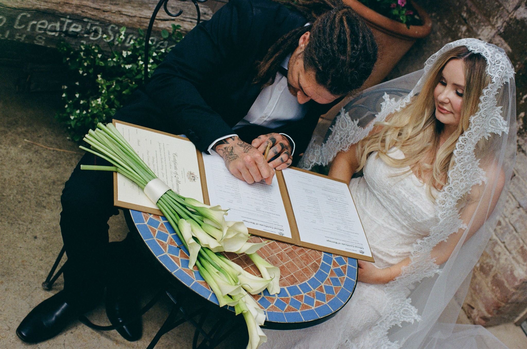 A man with tattoos signing a marriage certificate at a small round table, with a bouquet of calla lilies beside him, as a woman in a wedding dress and lace veil looks on smiling.