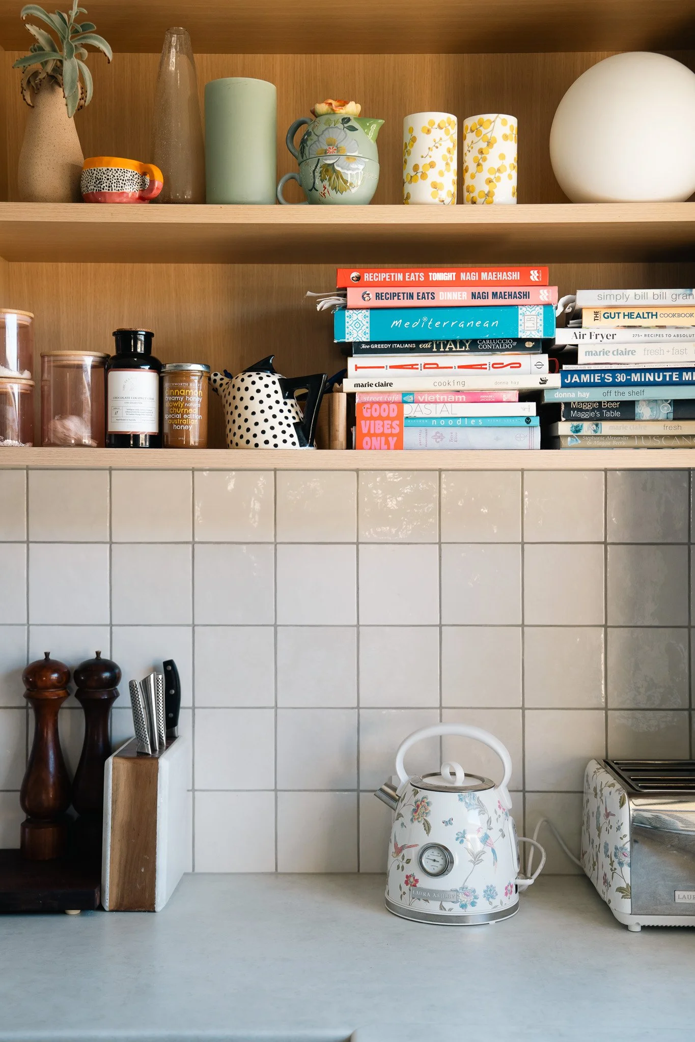 A kitchen shelf with various decorative vases and cups, and a stack of cookbooks. On the countertop, there are wooden pepper and salt shakers, a knife block, a floral-patterned kettle, and a floral-patterned toaster, Airbnb content.