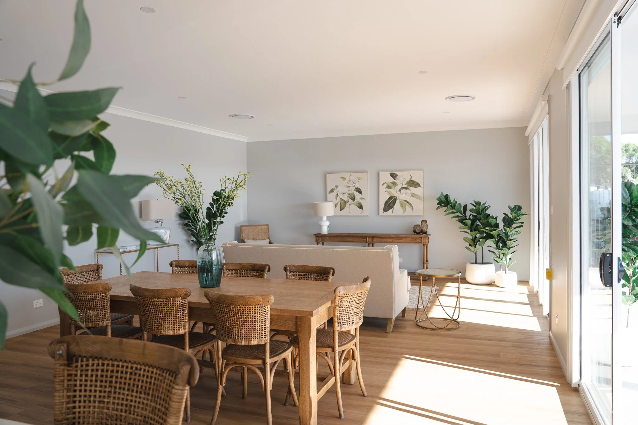 Bright living room with wooden dining table, wicker chairs, white sofa, and green plants near large sliding glass doors.