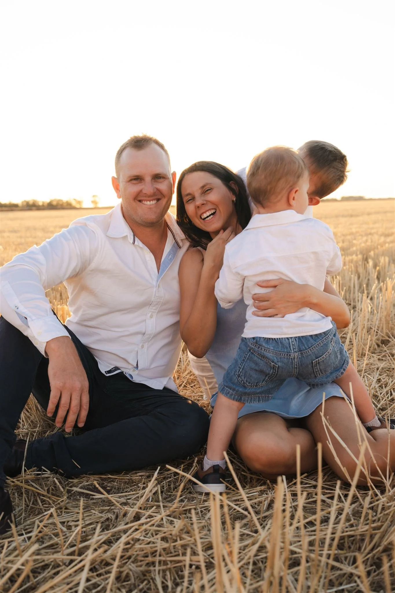 A family of four, sitting on a field during sunset, enjoying a joyful moment together. The father and mother are smiling while the young child is hugging the mother.