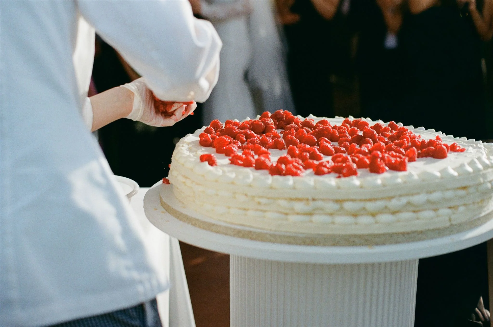 Wedding cake, Person in white gloves preparing a large white cake decorated with strawberries in front of a crowd.