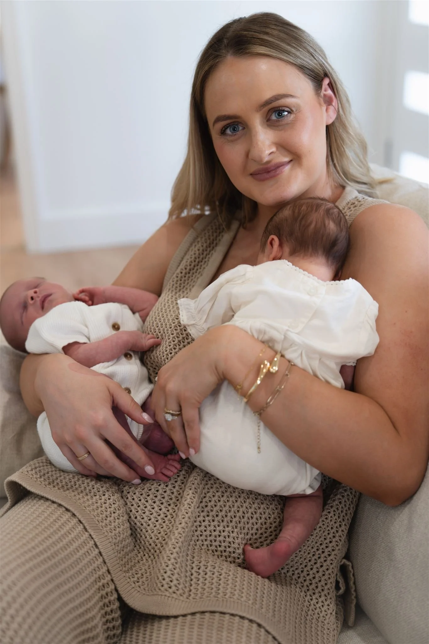 A woman holding two newborn babies, one in each arm, sitting on a beige couch in a well-lit room.