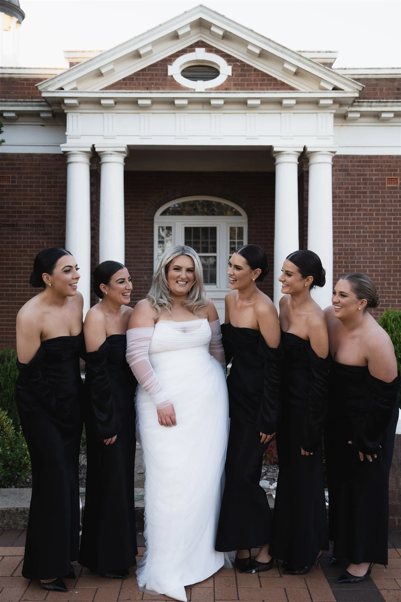 Group of women dressed in formal attire standing in front of a brick building with white columns, smiling and laughing.
