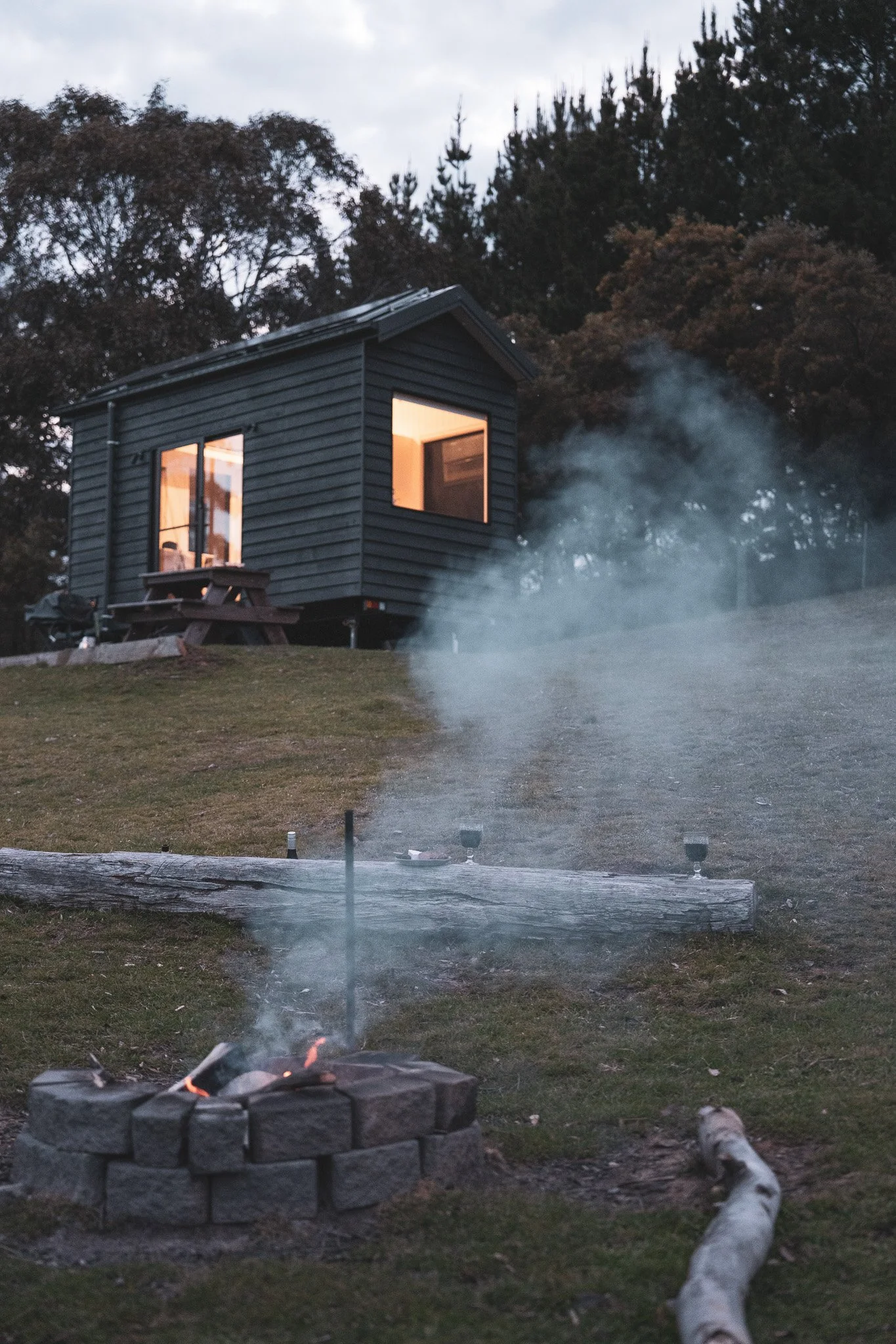 A small black house on a hill with lit interior windows, a picnic table outside, and a fire pit with smoke rising from it in the foreground, surrounded by grass and trees.
