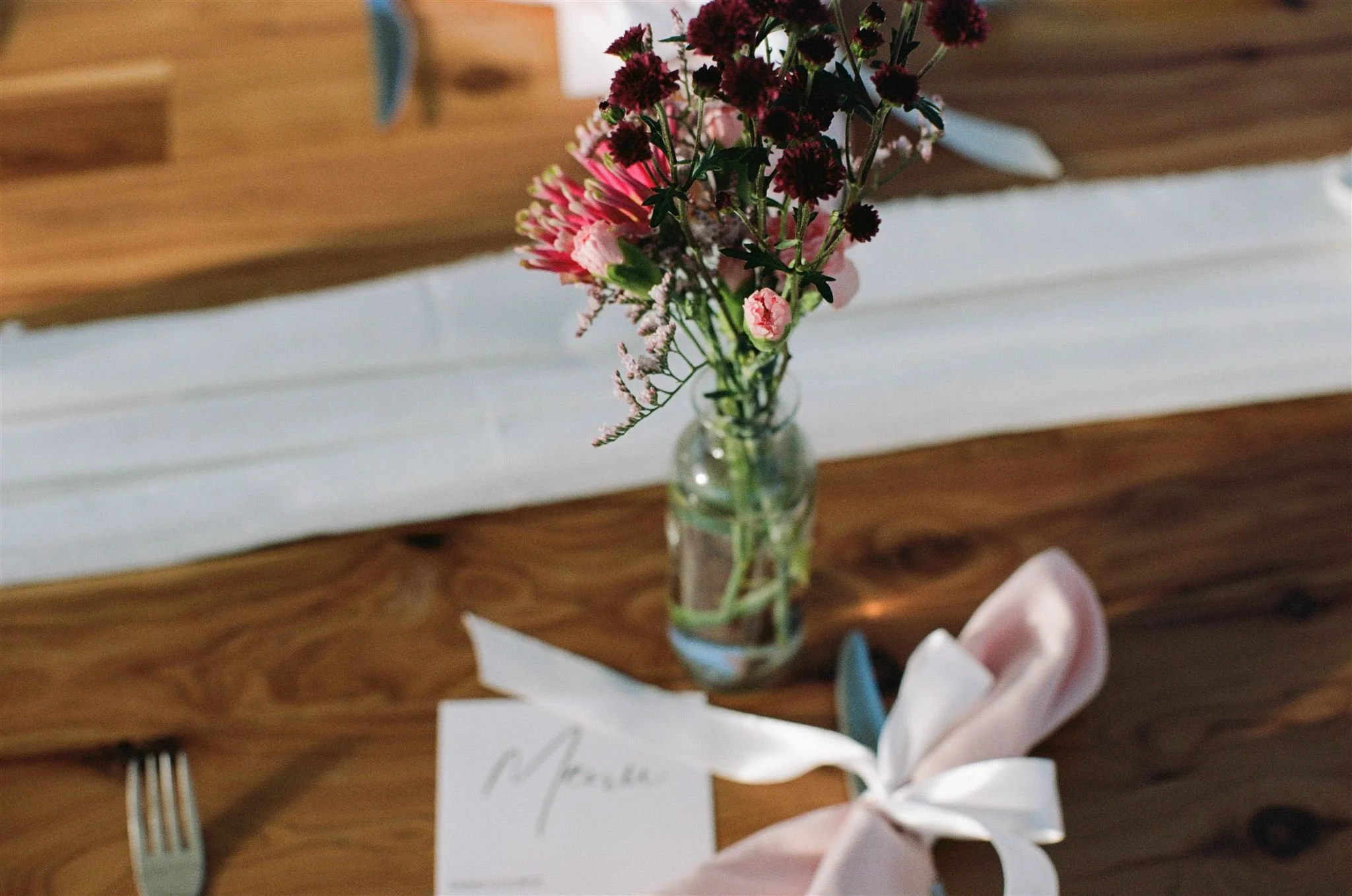 Griffith Golf Club Wedding. A small glass vase with pink and dark purple flowers on a wooden table. A white card with the word 'Menu' handwritten on it and a pink ribbon tied in a bow are also on the table.