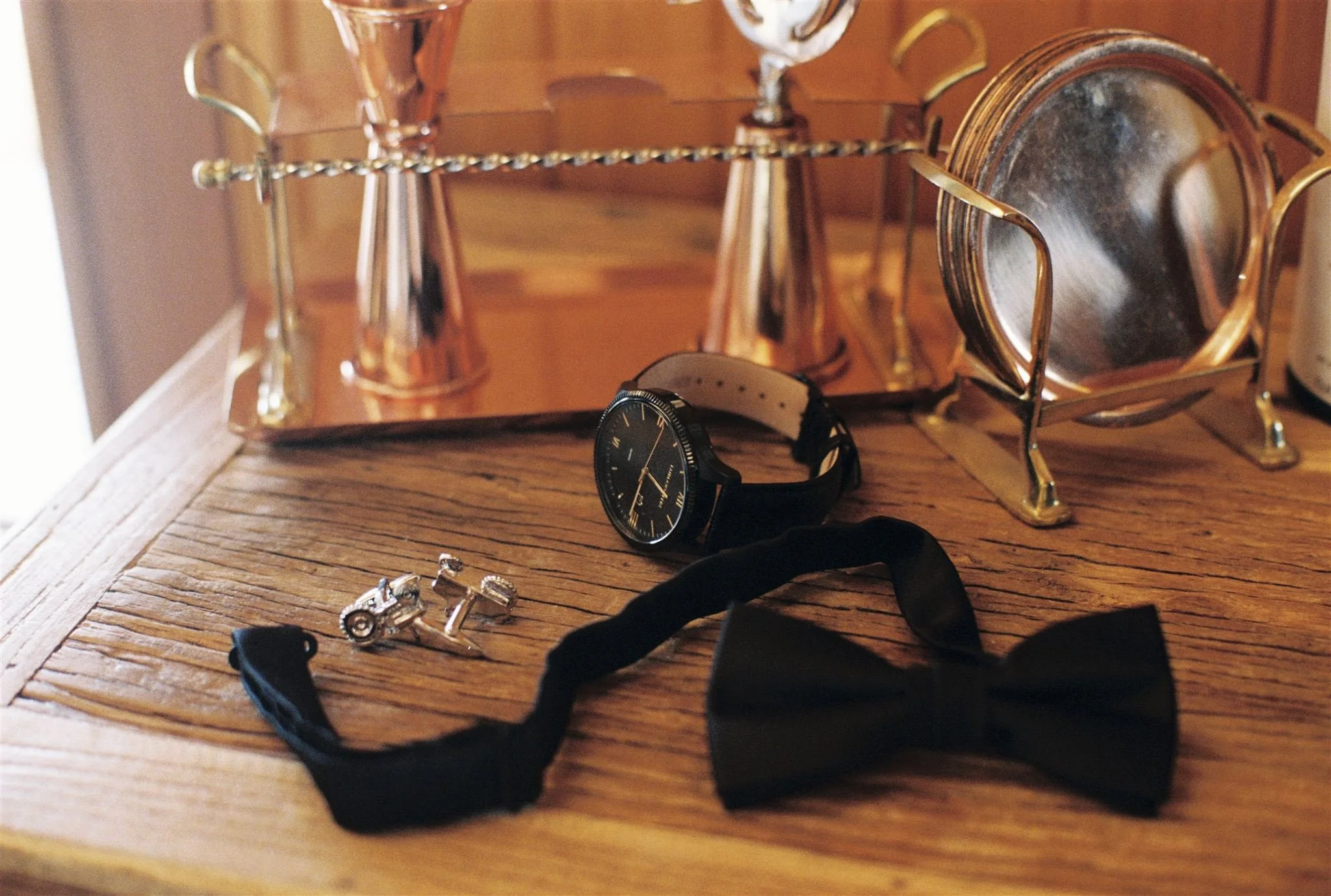 A wooden table displaying a black wristwatch with a white band, a black bow tie, a pair of silver motorcycle cufflinks, and a small black fabric item. In the background, a gold-colored tray with a mirror and two silver trophies.