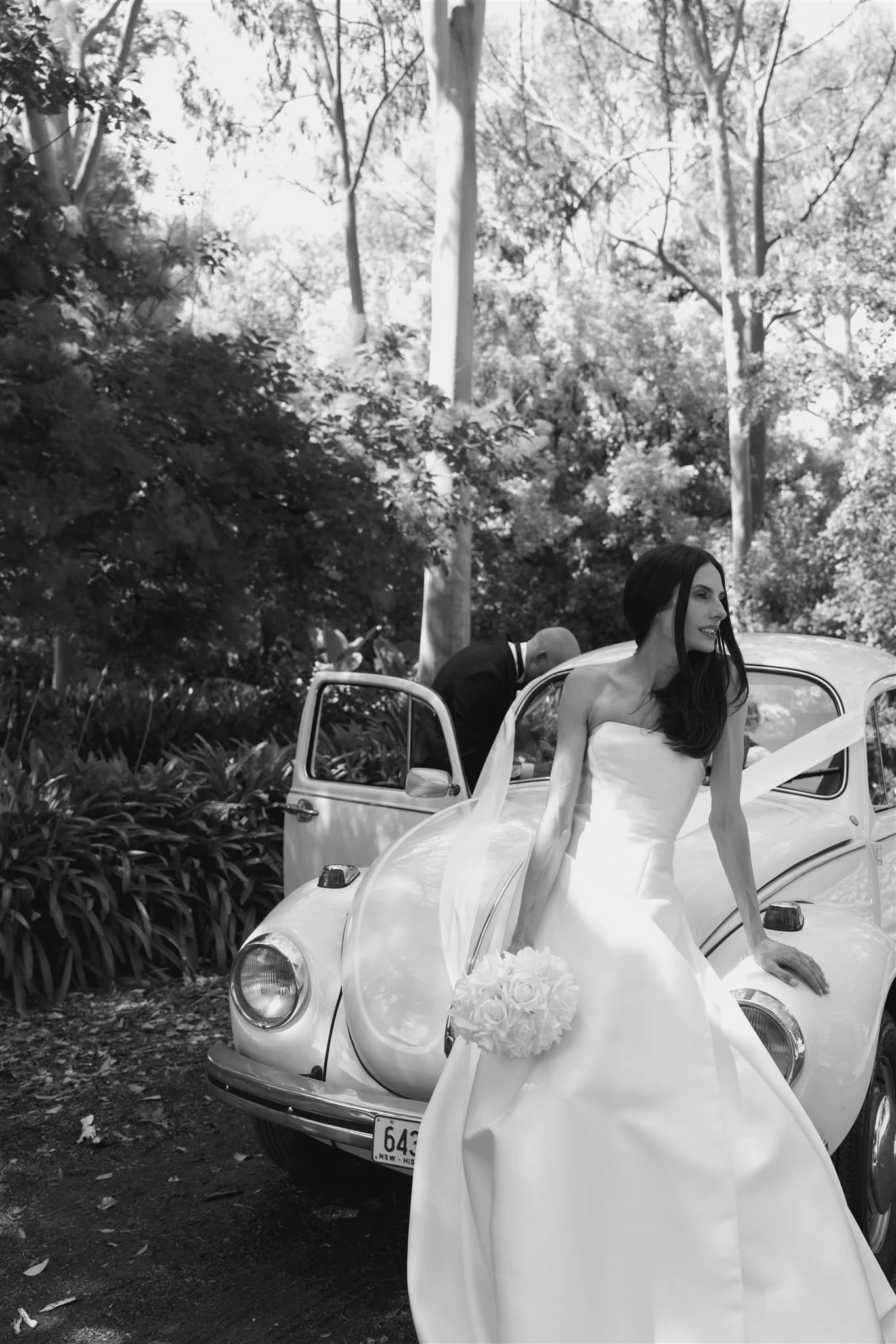A bride in a strapless wedding dress holding a bouquet of roses, standing beside a vintage car, with a man in a suit behind her, in a wooded outdoor setting.