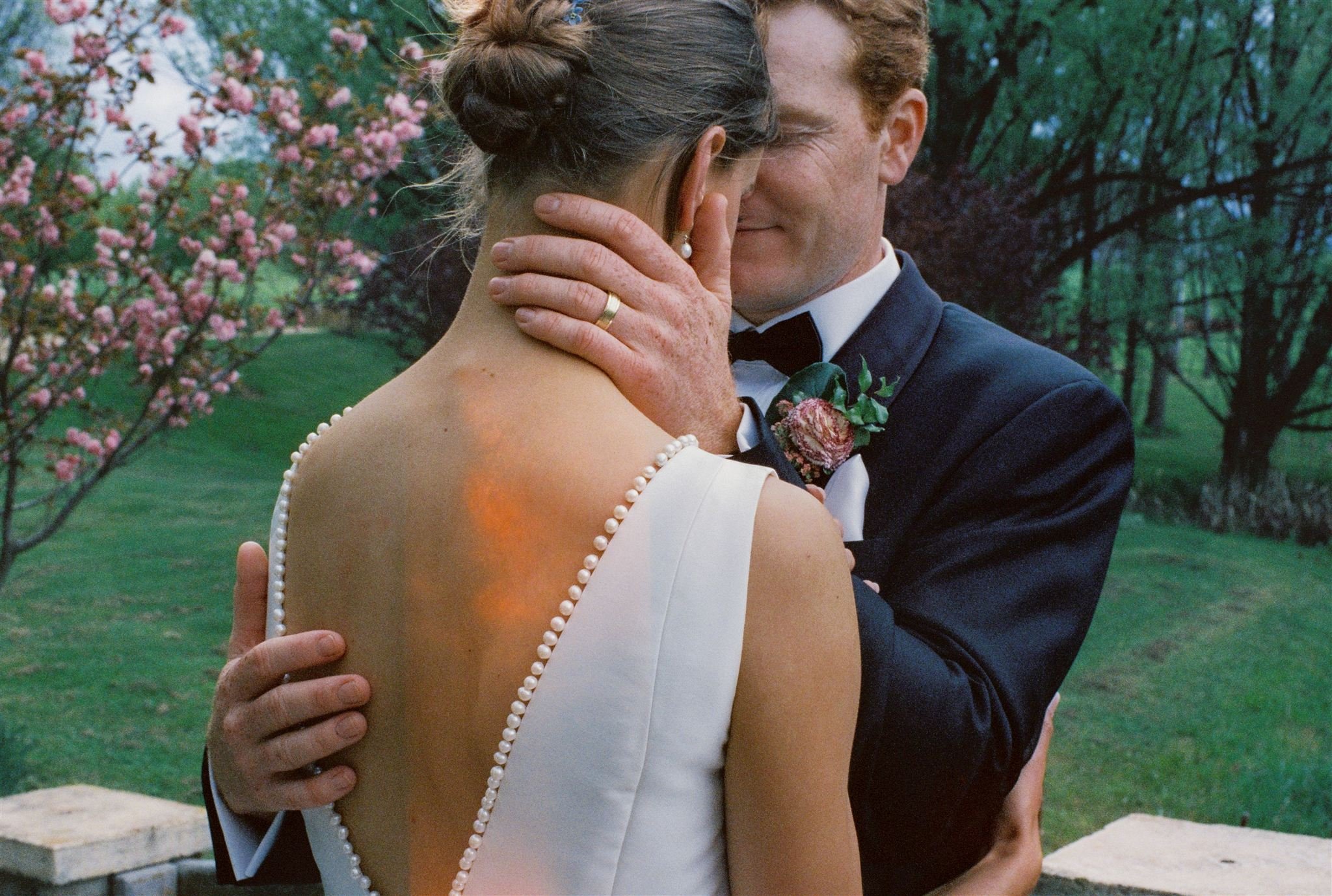 Waldara Farm Wedding. A bride and groom share a close, intimate moment outdoors on their wedding day, with the bride's back to the camera and the groom gently holding her face, surrounded by pink blossoms and green trees.