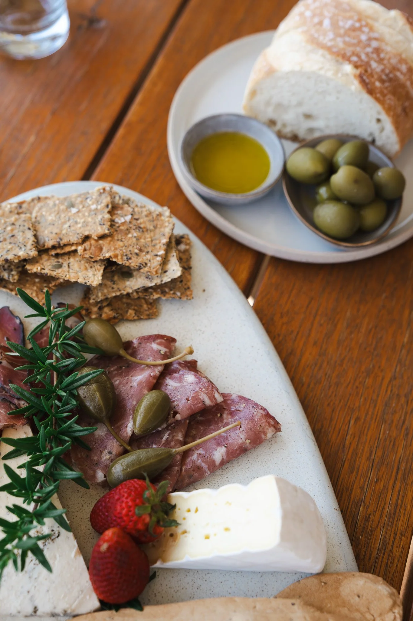 A cheese and charcuterie board with grapes, strawberries, crackers, and herbs, with a side of bread, olives, and olive oil on a wooden table.