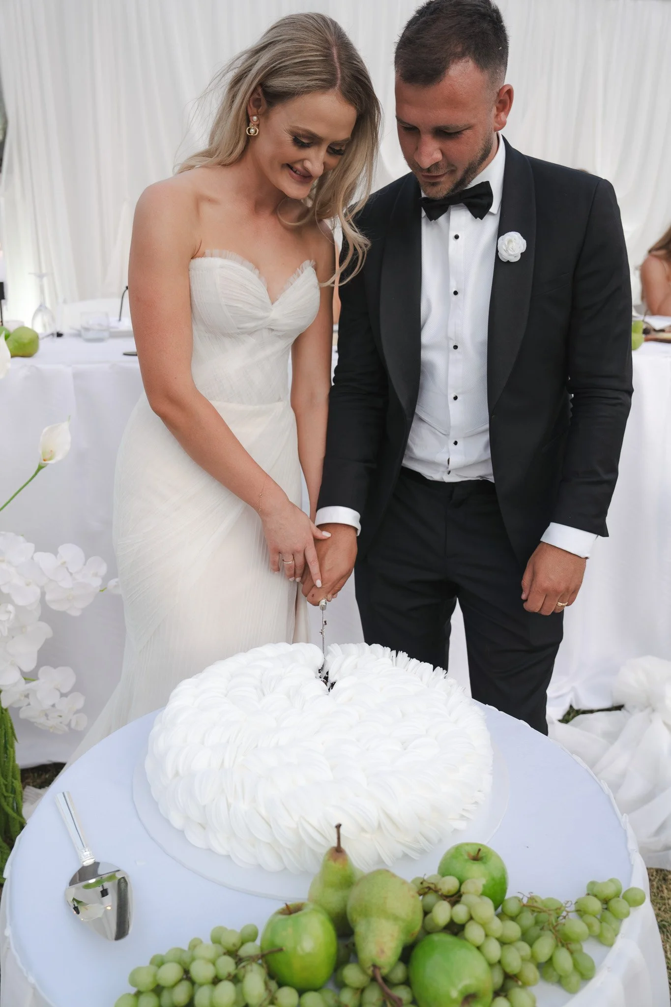 A bride and groom cutting a wedding cake together at their wedding reception. The bride is wearing a white strapless gown, and the groom is in a black tuxedo.