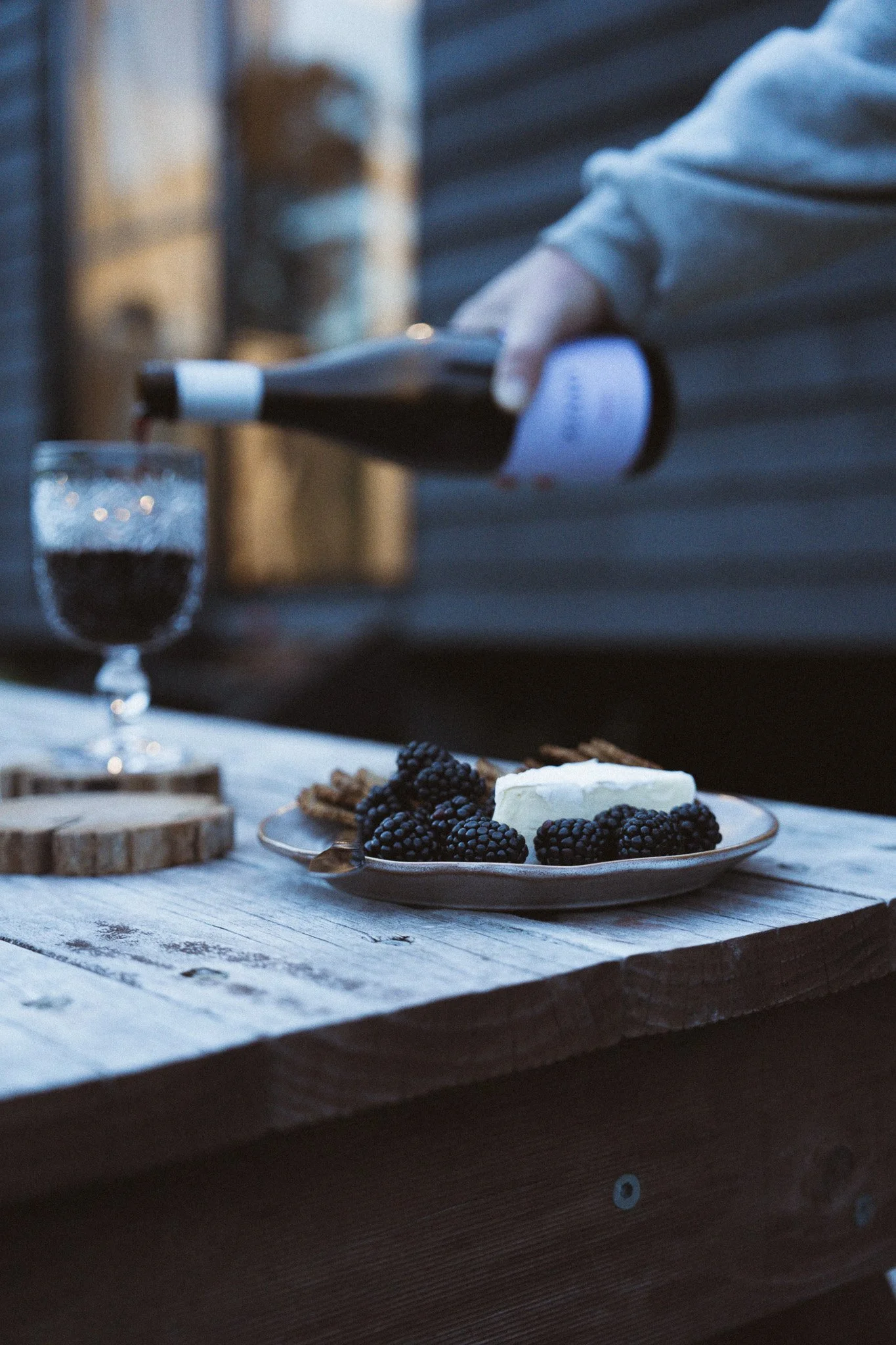 A person is pouring red wine into a glass, with a cheese wedge and blackberries on a plate in the foreground, on a rustic wooden table outdoors.