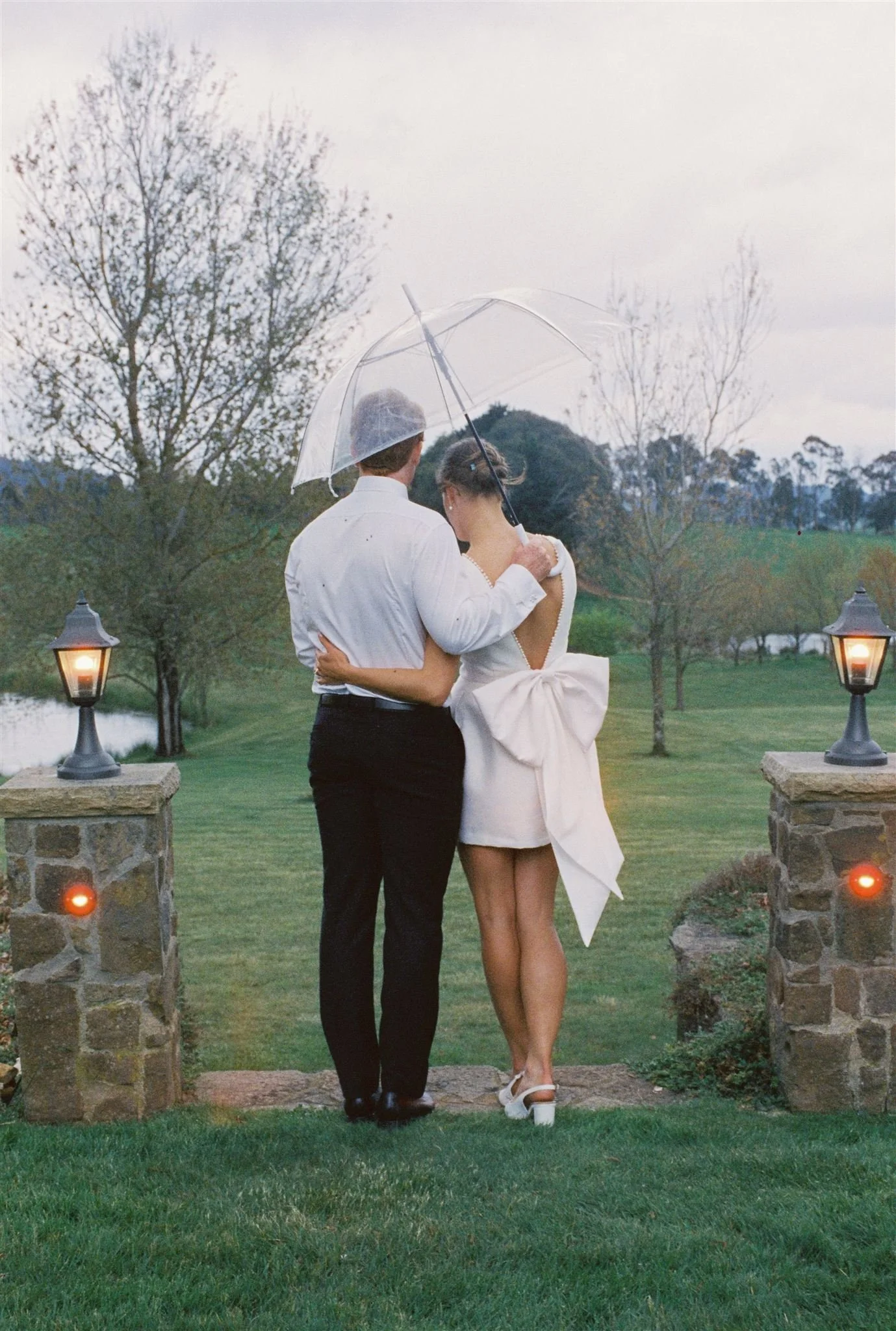 A couple standing outdoors on a grassy area during daylight, sharing an umbrella. The man wears a white shirt and black pants, while the woman wears a white dress with a large bow at the back. There are stone pillars with lanterns on top on either si