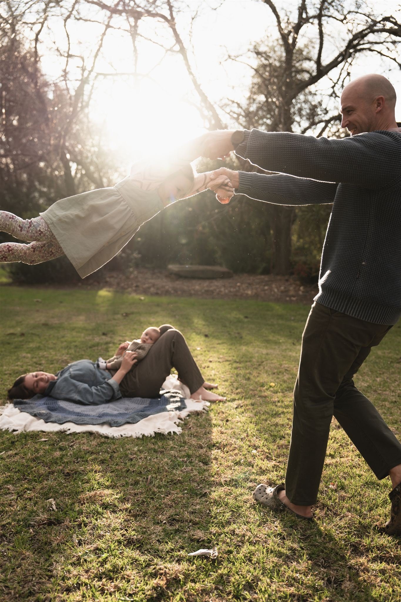 A family enjoying a playful moment outside on a sunny day. An adult man is holding hands with a young girl who is being lifted off the ground by him, while a woman and baby are lying on blankets on the grass, watching and smiling.