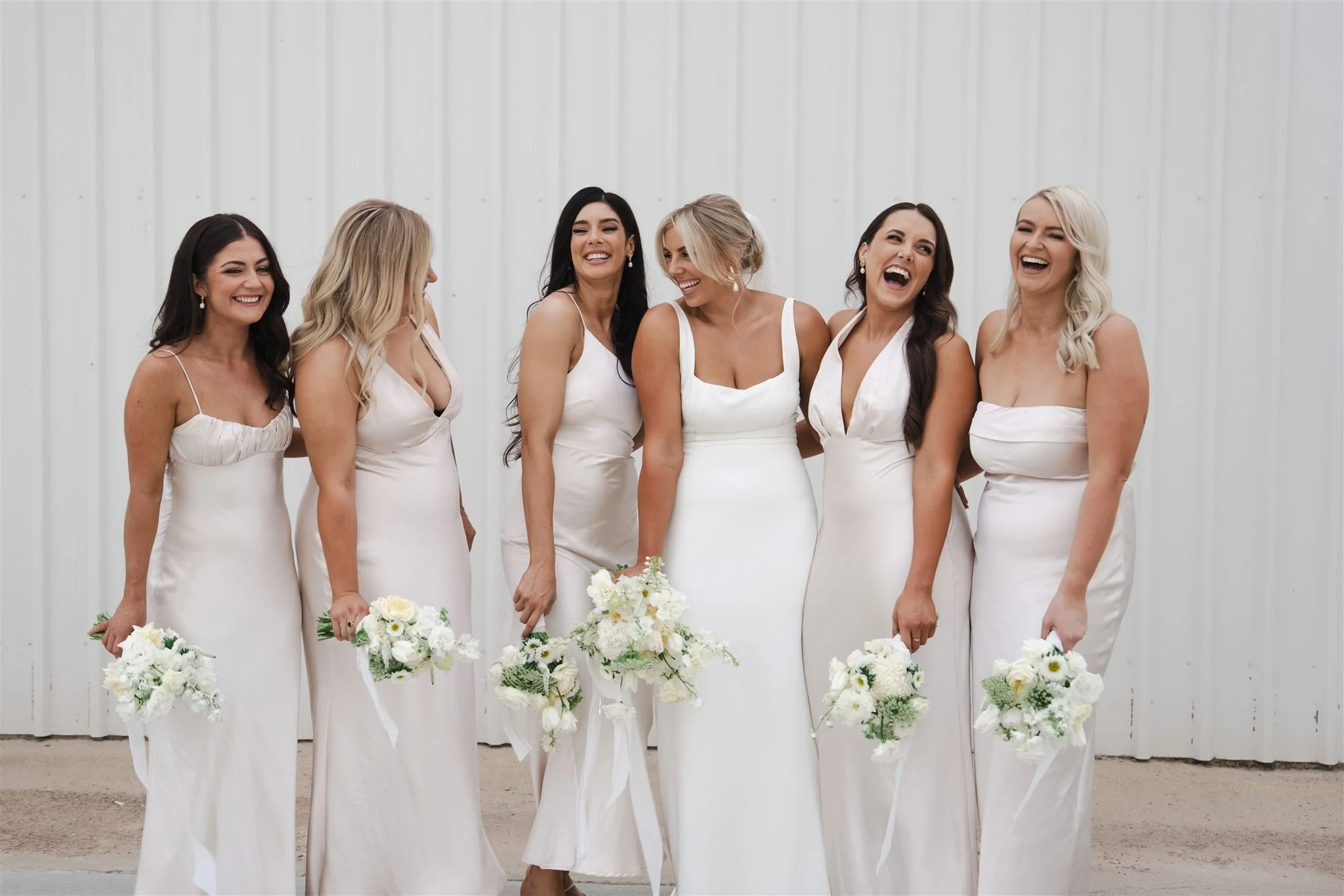 Group of six women in white dresses holding white flower bouquets, standing against a white wall, smiling and laughing.