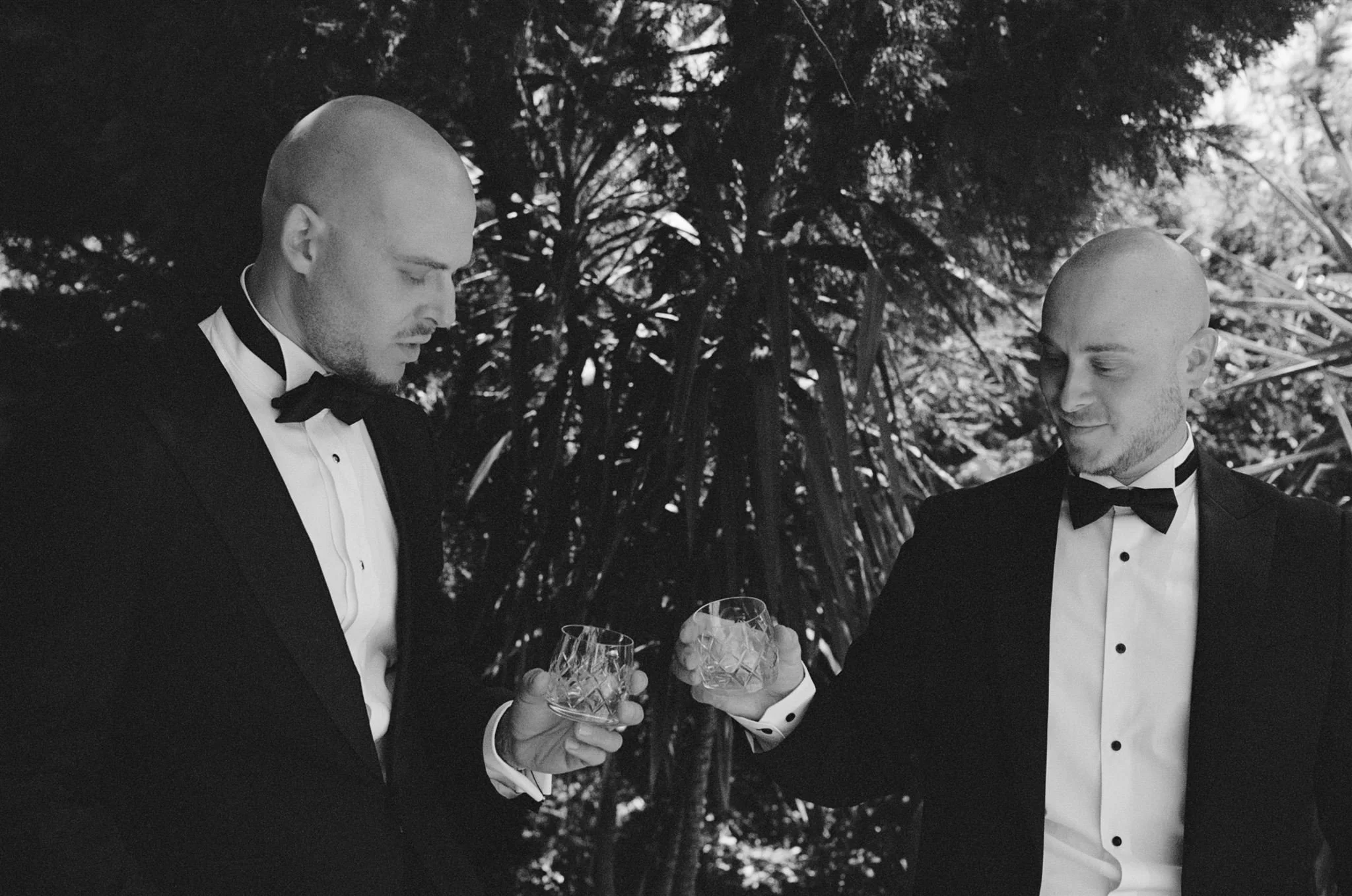 Two men in tuxedos with bow ties, bald and bearded, toasting with glasses in an outdoor setting with trees in the background.