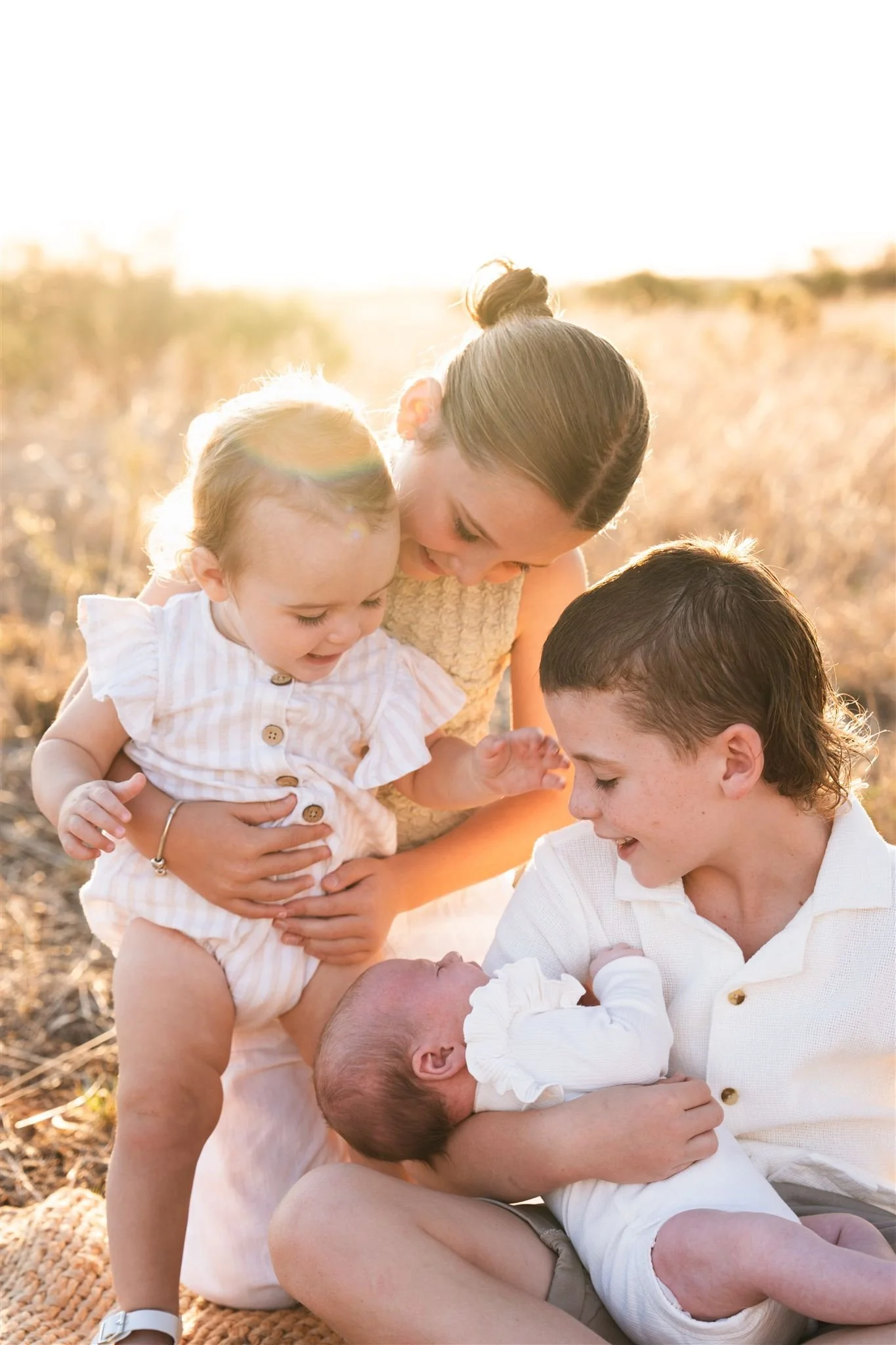 A family of five, including two young children and a baby, sitting outside on a sunny day in a field, sharing a tender moment together.