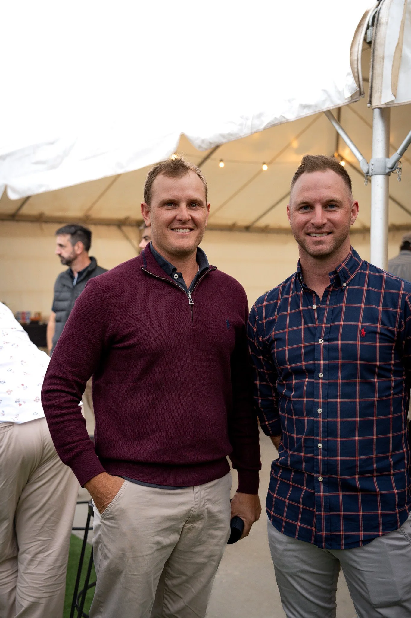 Two men smiling at an outdoor event under a tent, with other people in the background.