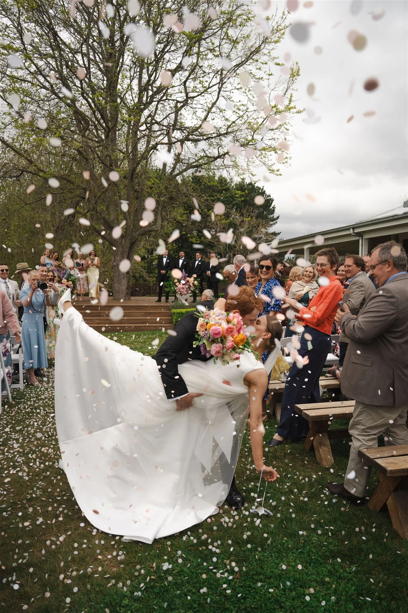 A bride and groom sharing a kiss while the groom dips the bride during a wedding celebration outdoors, with guests throwing confetti.