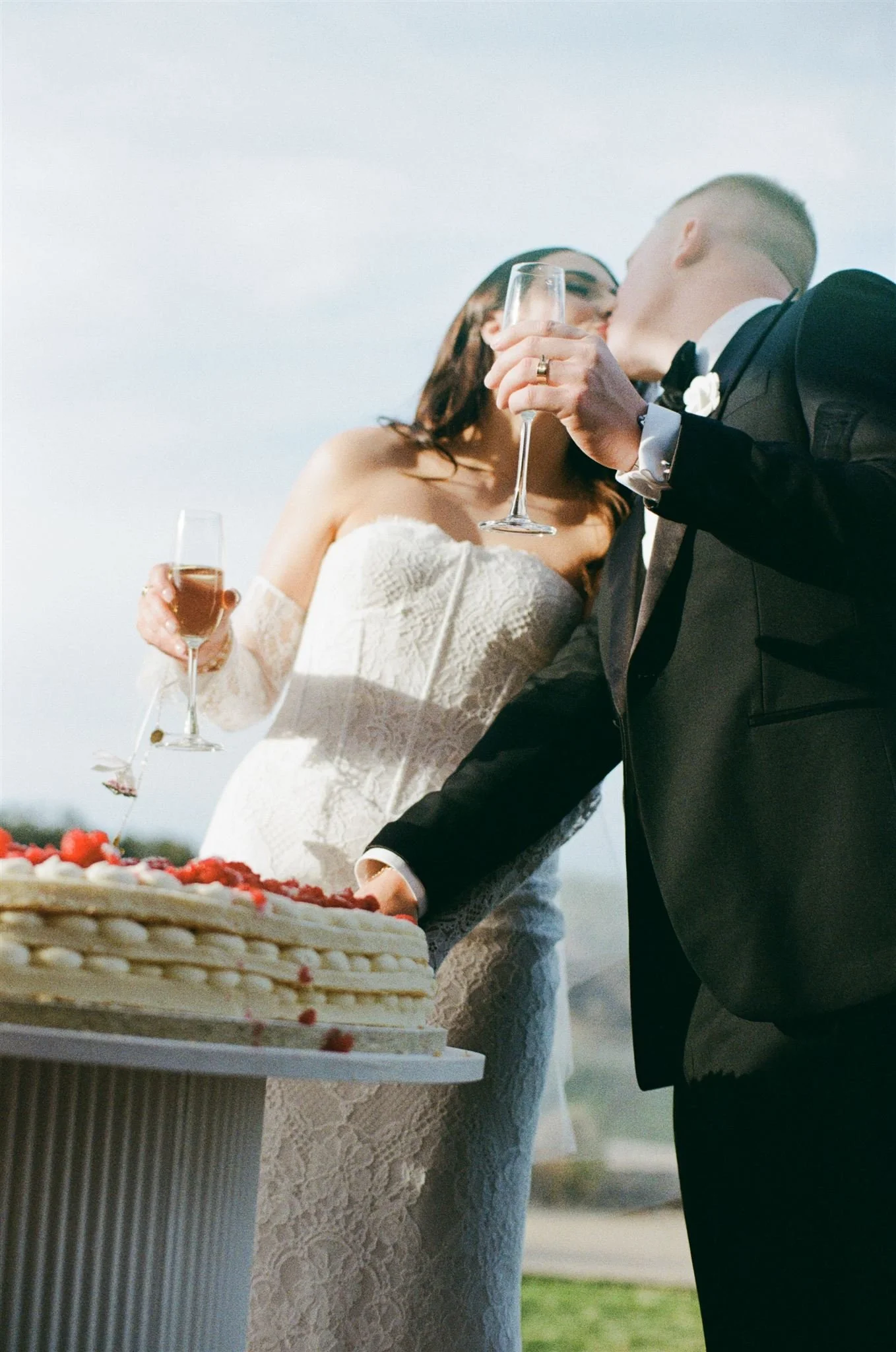 Whitton Malt House wedding photography. Bride and groom kissing during their wedding reception, lifting glasses of champagne, with a wedding cake on a table
