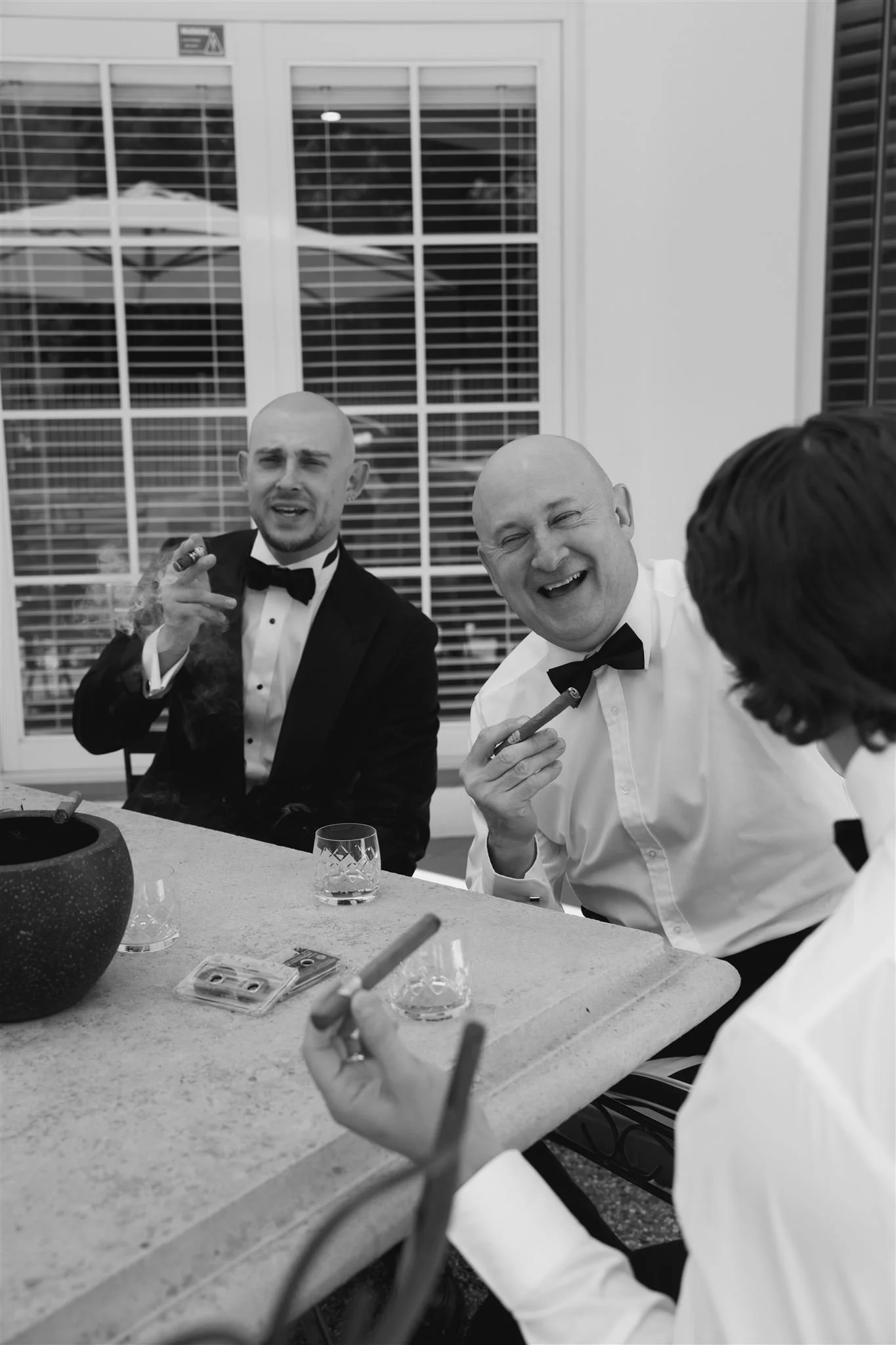 Three men dressed in tuxedos and bow ties sitting at a table, smiling and talking, with cigars in hand during a social gathering in a bright room with large windows and blinds.