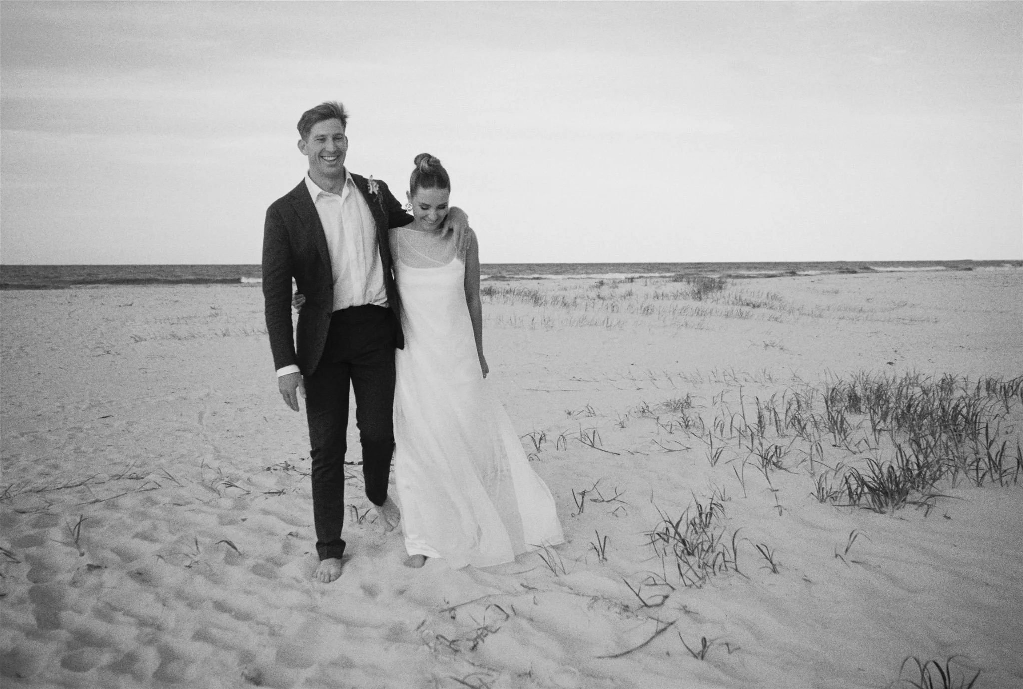 Australian coastal photography, east coast. black and white photo of a smiling man and woman walking barefoot on a sandy beach. The man is wearing a suit, and the woman is in a white dress. She has her arm around his shoulder as they walk together.