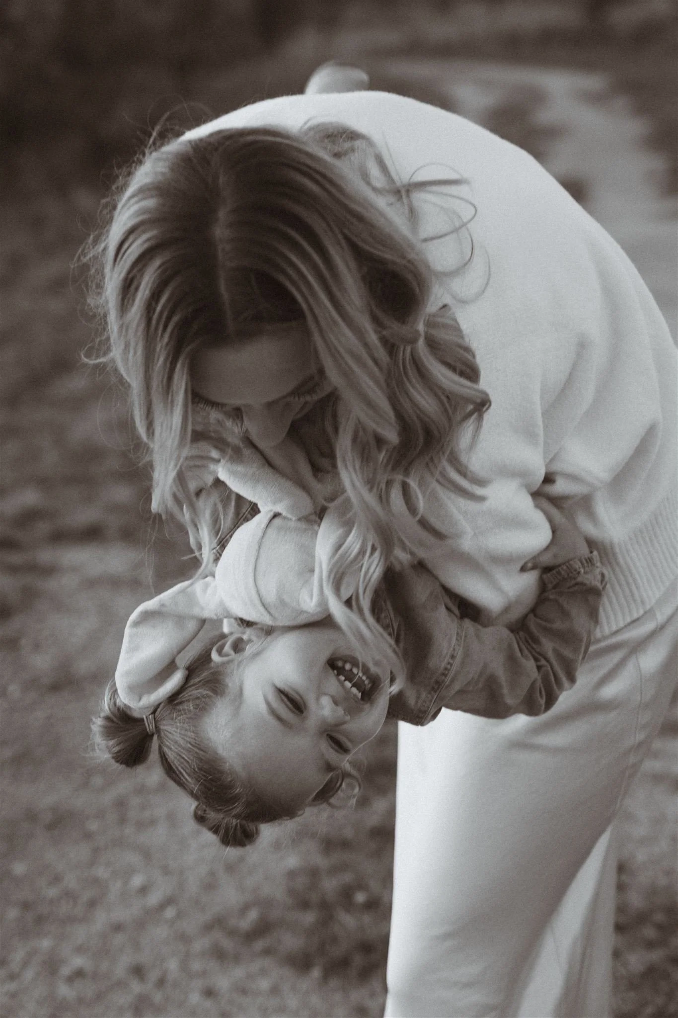 A woman is playfully holding a young girl upside down, both laughing and enjoying a moment outdoors.