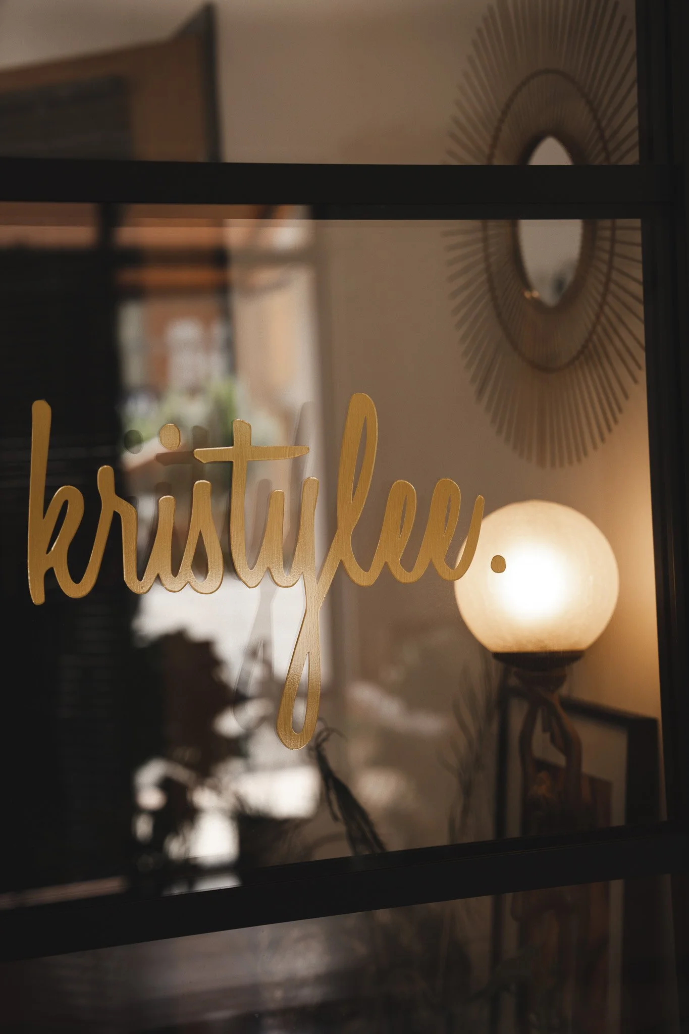 A glass window with golden cursive text that says 'krystalu', reflecting a warmly lit interior with a round white lamp and decorative wall mirror, jewellery, content.