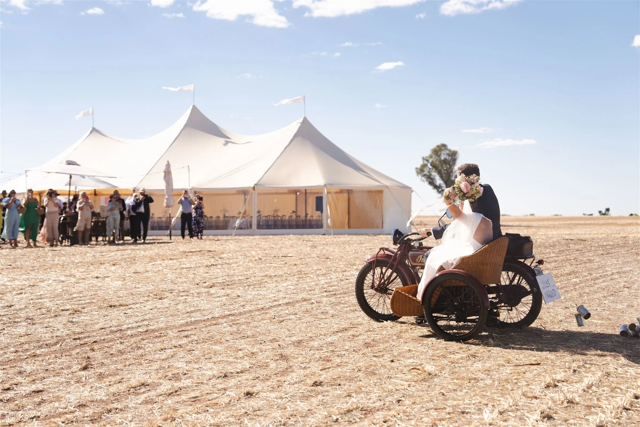 A bride and groom celebrating their wedding on a tricycle in an open field with a large white wedding tent in the background, bright blue sky, and guests nearby.