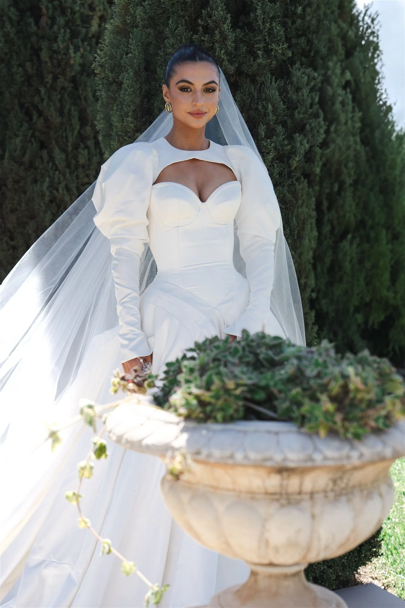 bride, A woman wearing a white wedding gown with puffed sleeves, a sweetheart neckline, a veil, and gold earrings, standing outdoors in front of a green hedge.