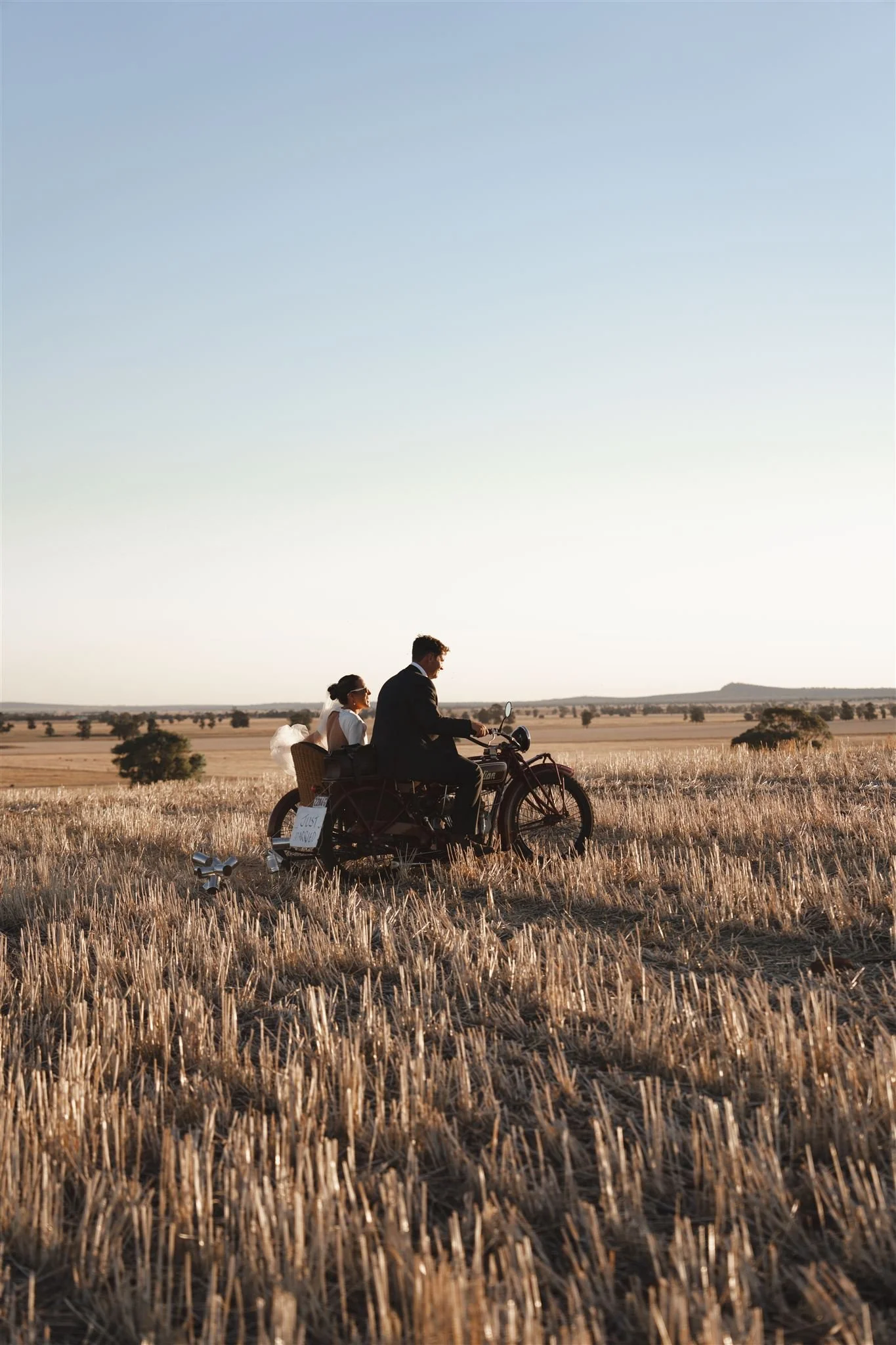 bride and groom portrait, country wedding, A couple riding a motorcycle with a sidecar through a golden wheat field during sunset. The man is driving, and the woman is sitting in the sidecar.